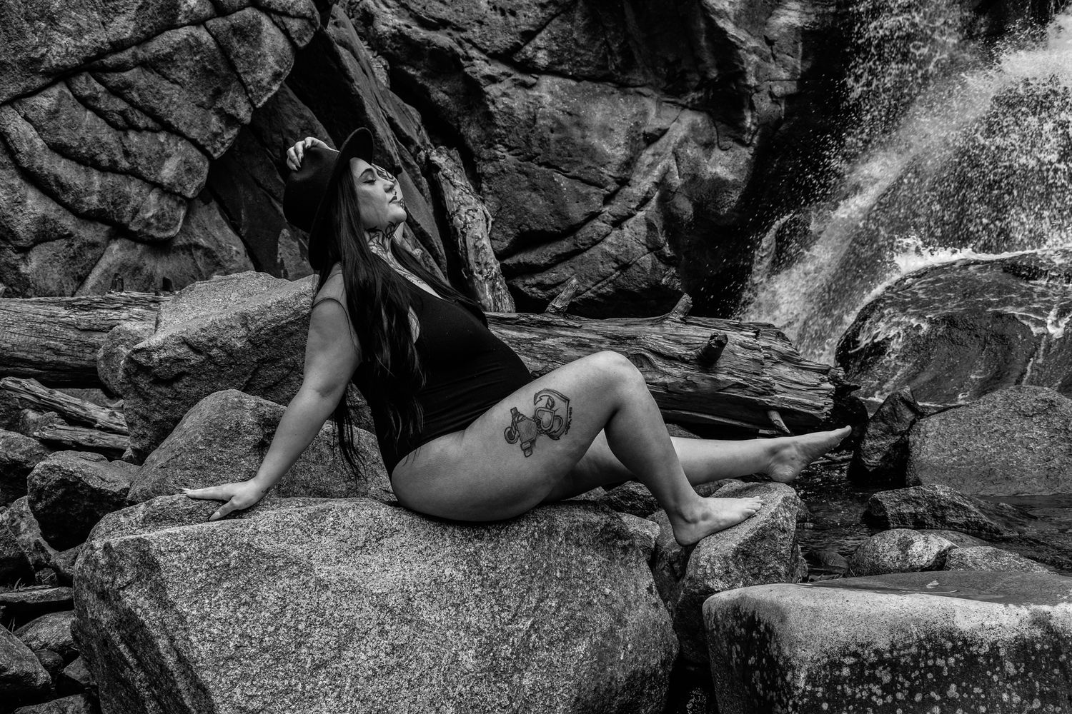 Black and white photo of tattooed woman in swimsuit reclining on rocks by waterfall.