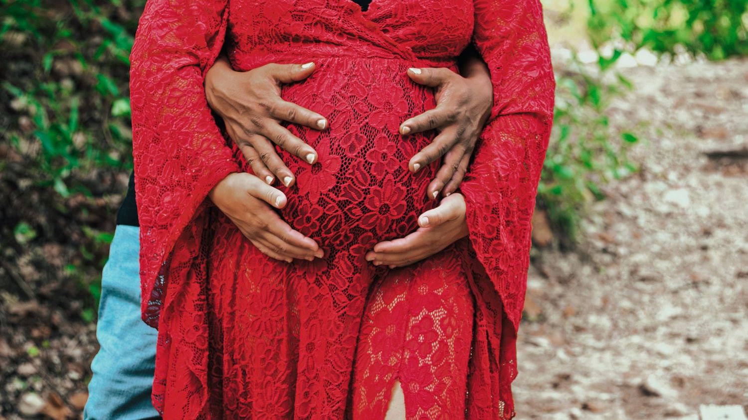 Close-up of two pairs of hands cradling a pregnant belly in a red lace maternity dress outdoors.