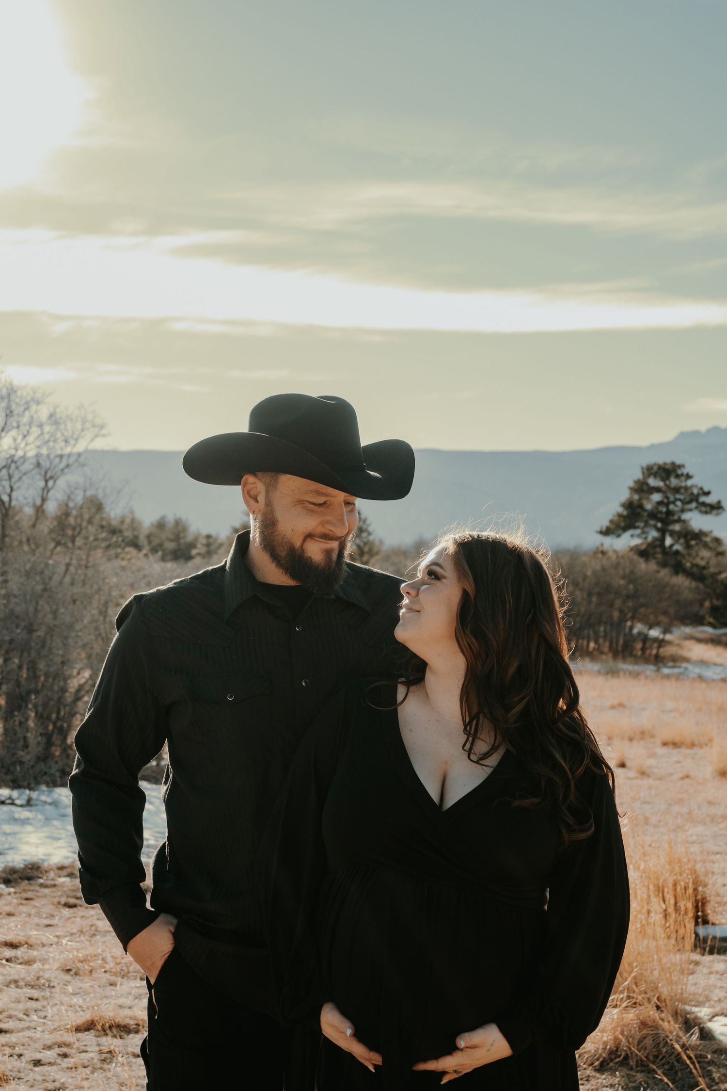 Man in black cowboy hat and woman in black dress sharing a loving look during winter maternity session.