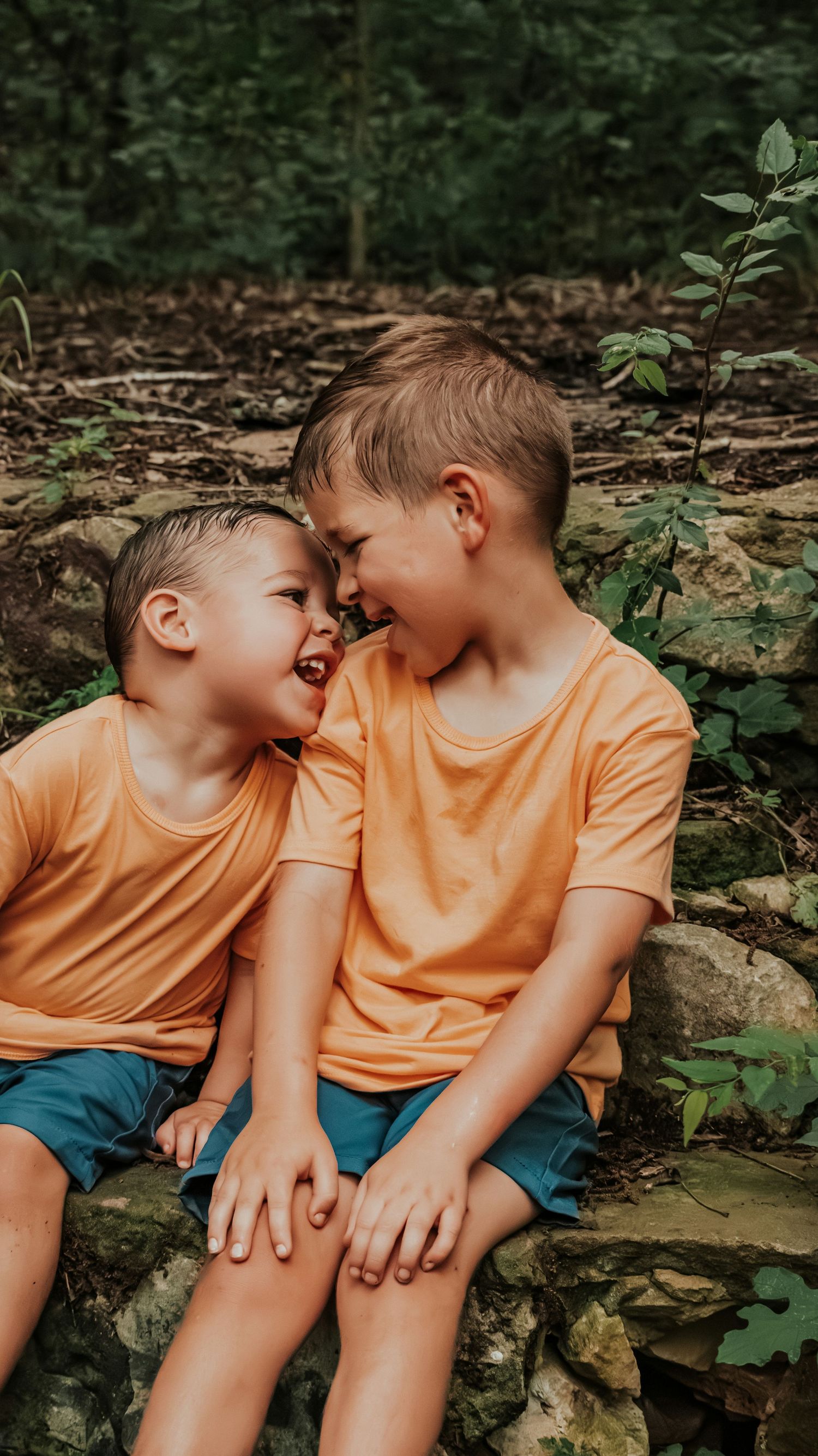 Two young brothers in matching orange shirts laugh together while sitting on a stone wall outdoors.