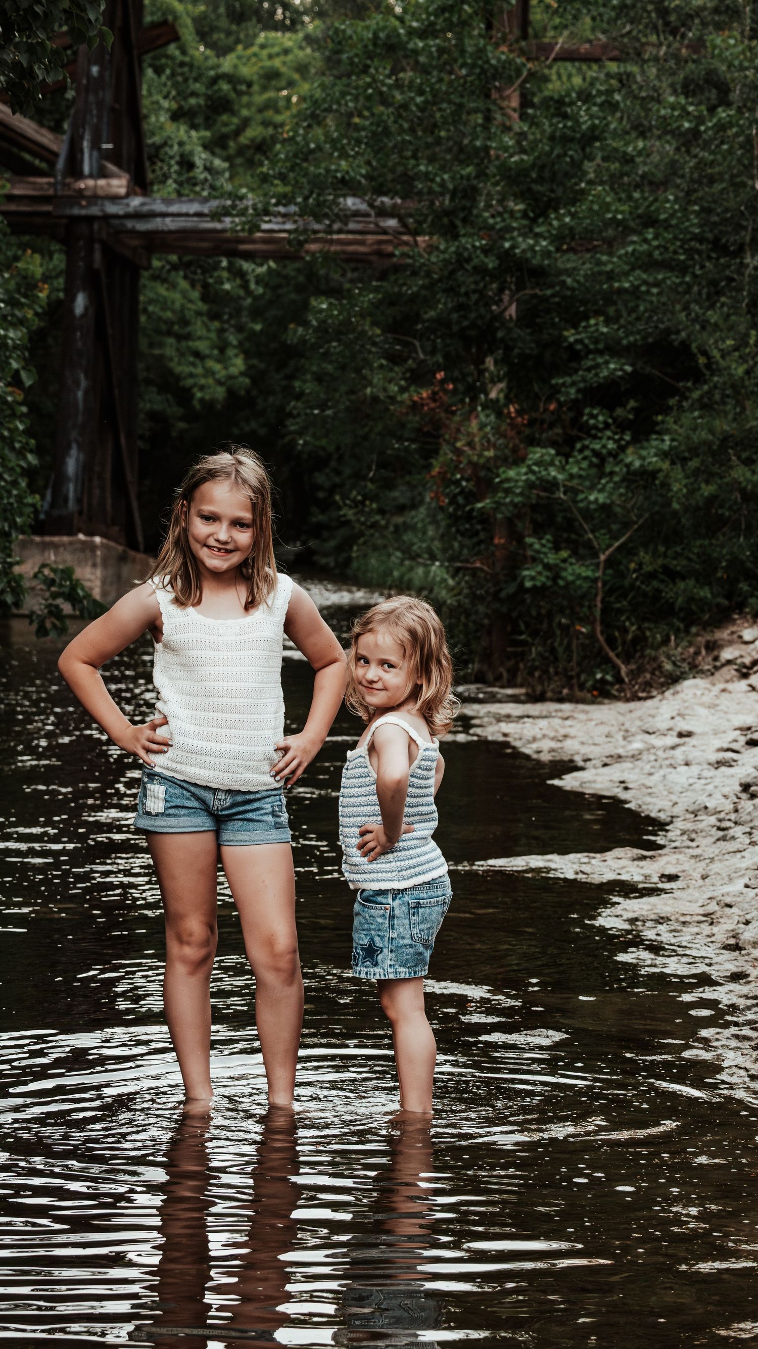 Two young sisters in white tops and denim shorts posing together in a shallow river.