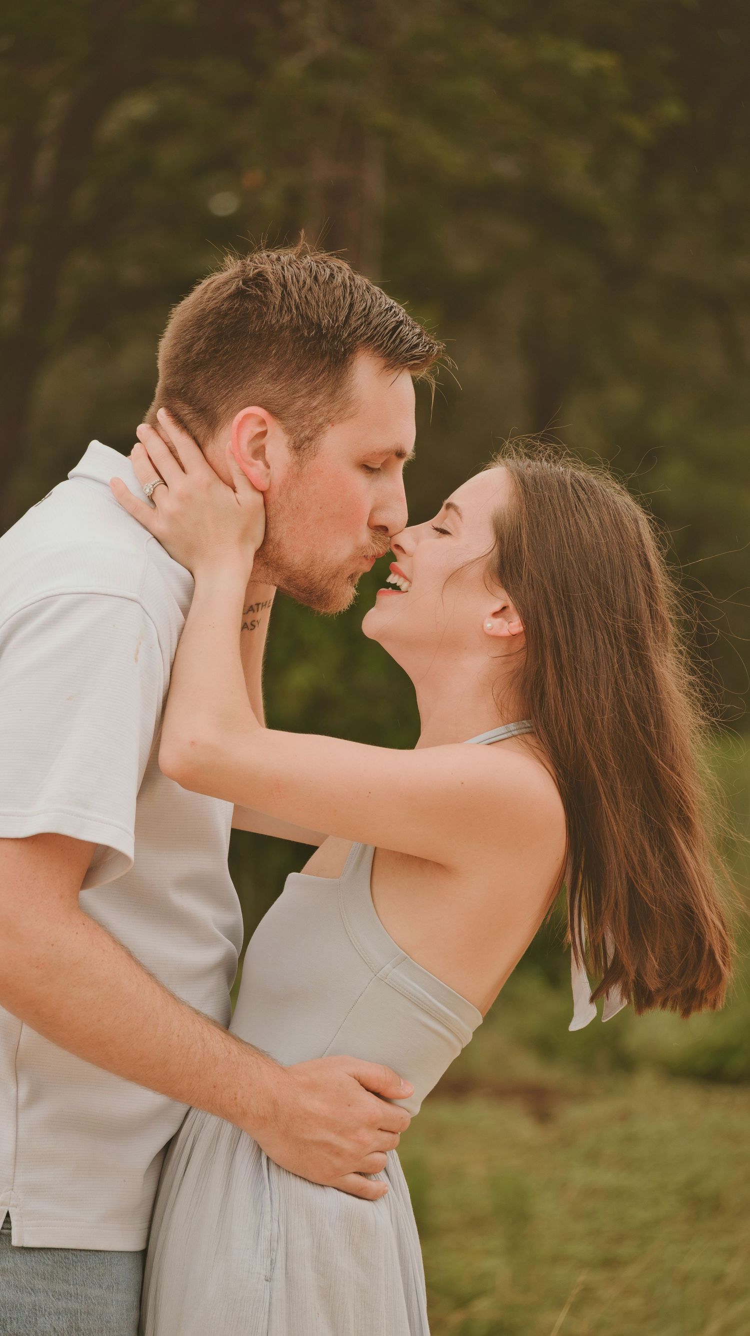 A couple shares a romantic kiss outdoors with lush green trees in the background for a family session.
