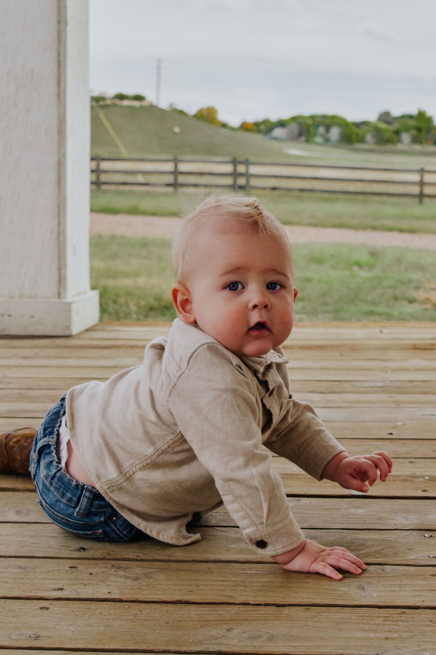 Blonde baby boy in beige shirt crawling on wooden deck with farm field in background.