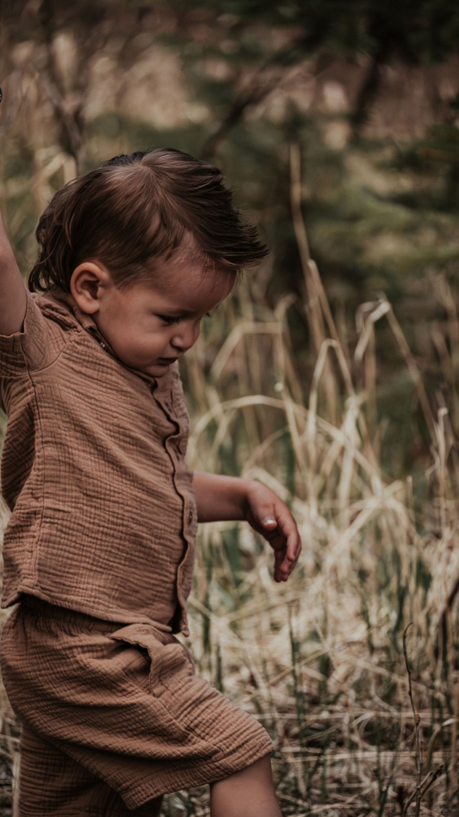 Adorable toddler boy in brown linen outfit exploring nature in a grassy outdoor setting.