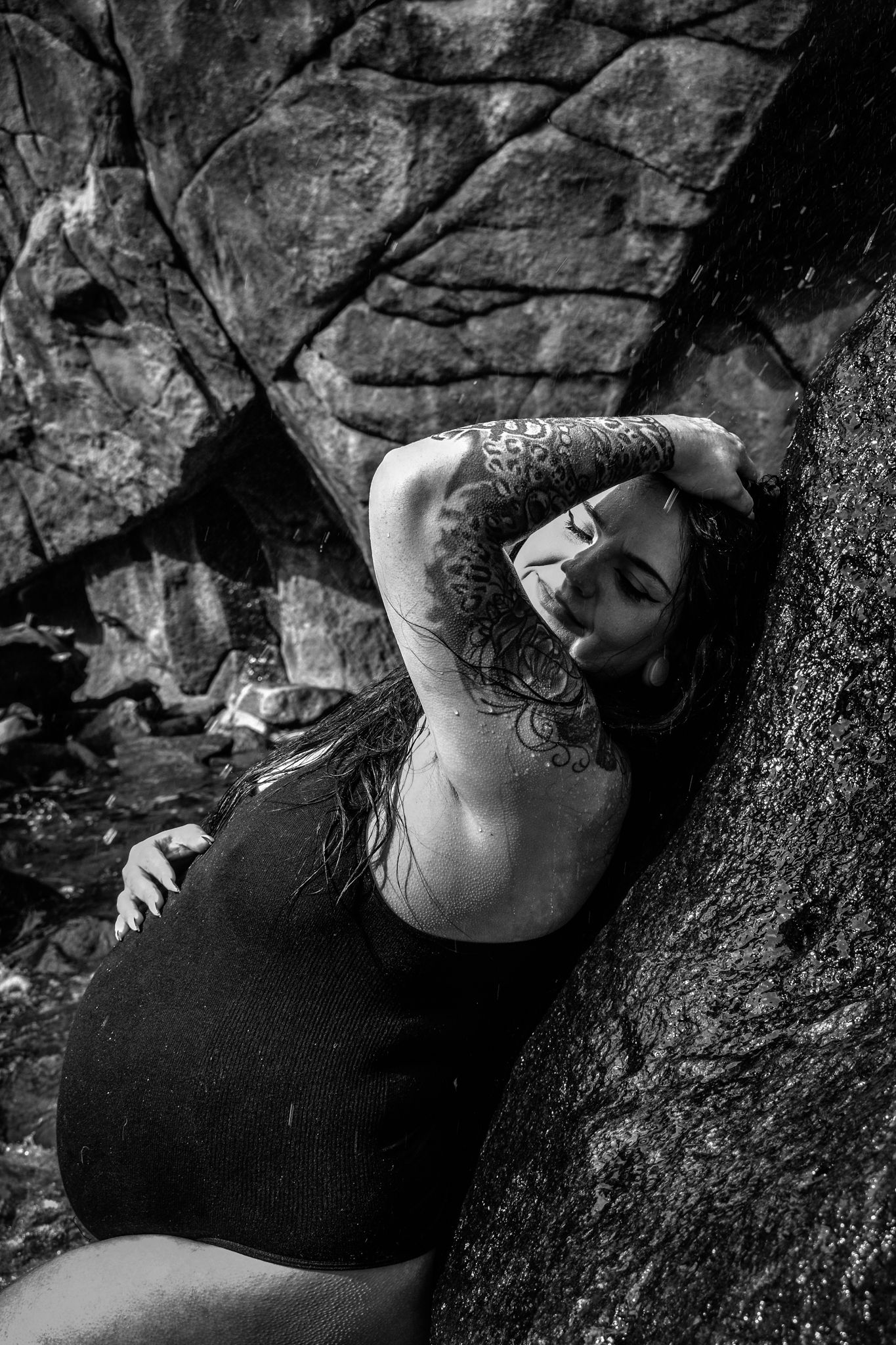 Black and white photo of tattooed woman in black swimsuit leaning against large rock outdoors.