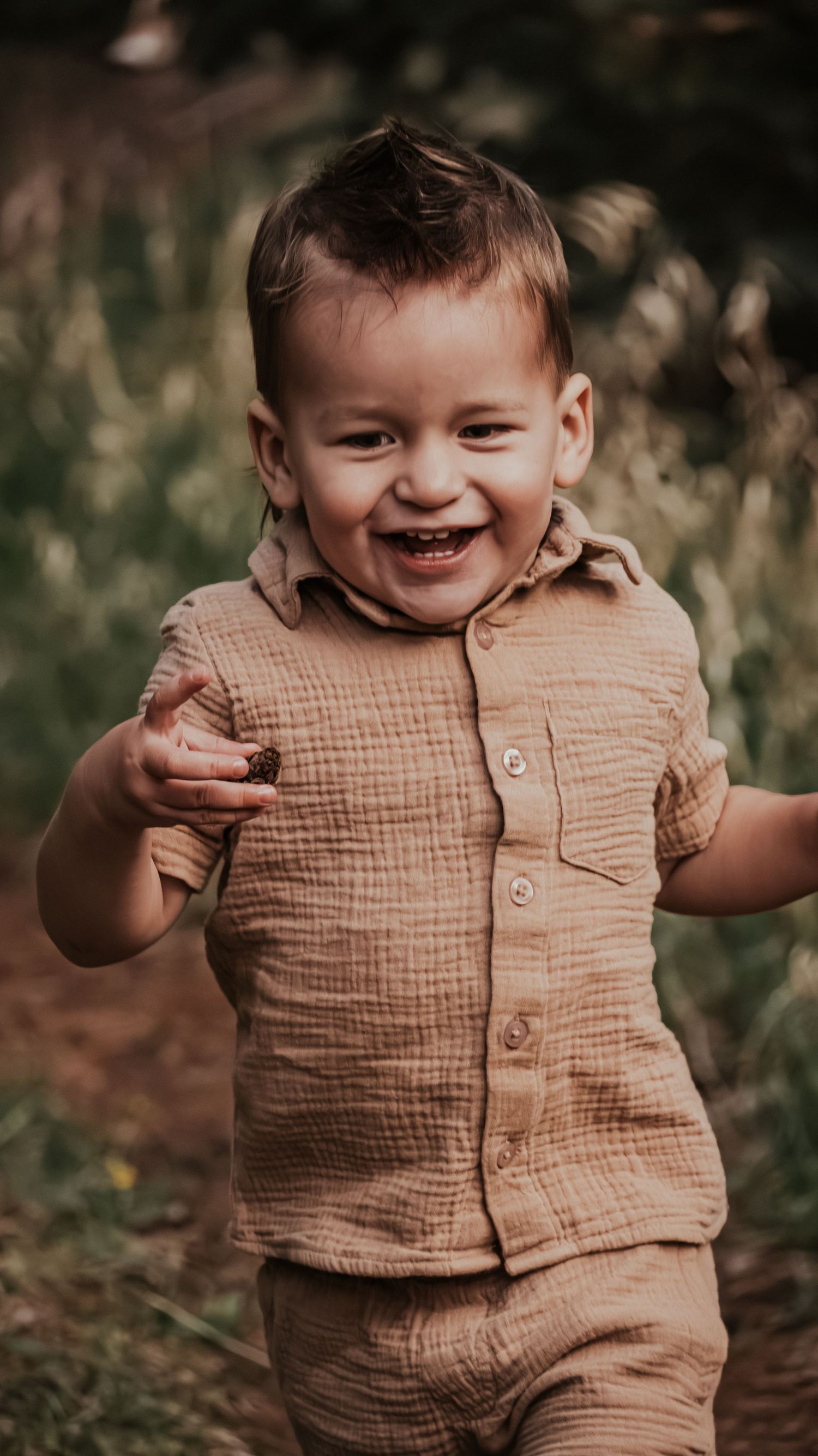 A cheerful toddler boy in a tan linen outfit runs and smiles while holding something in his hand.