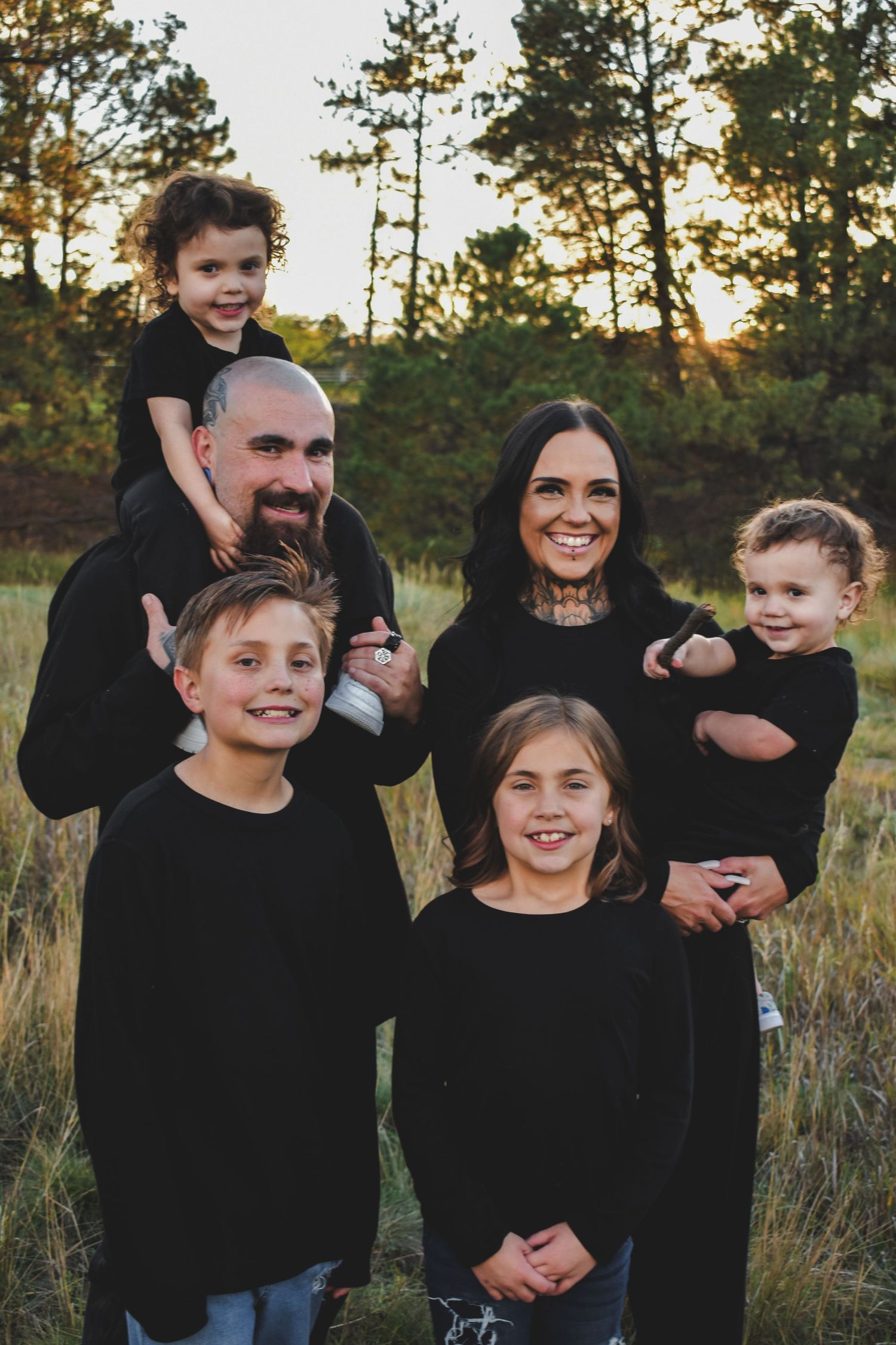 A joyful family of six poses outdoors in matching black outfits among tall grass and pine trees at golden hour.
