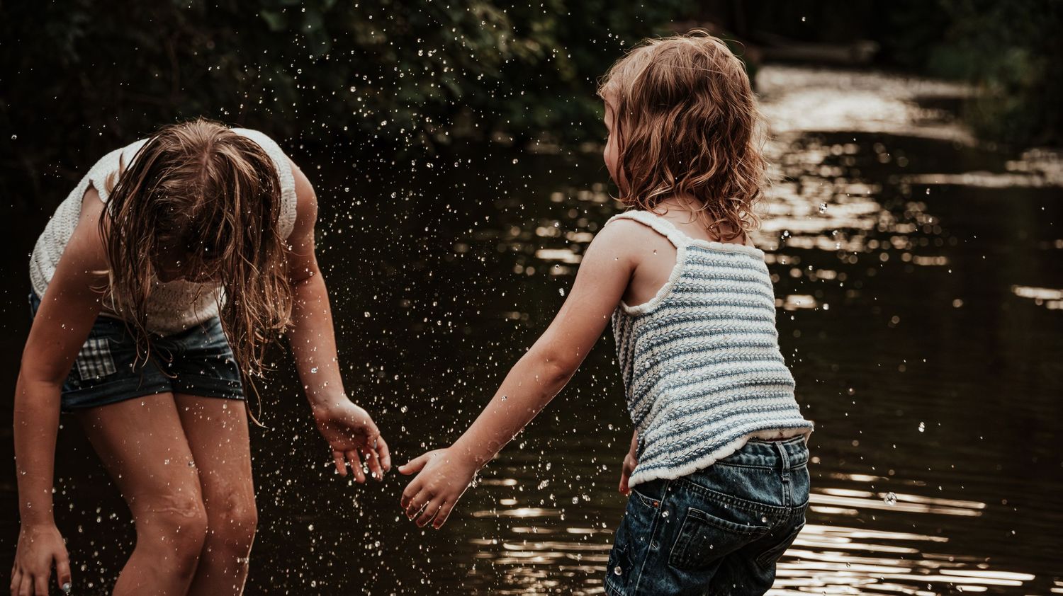 Two children holding hands splashing in a river with water droplets caught in motion.