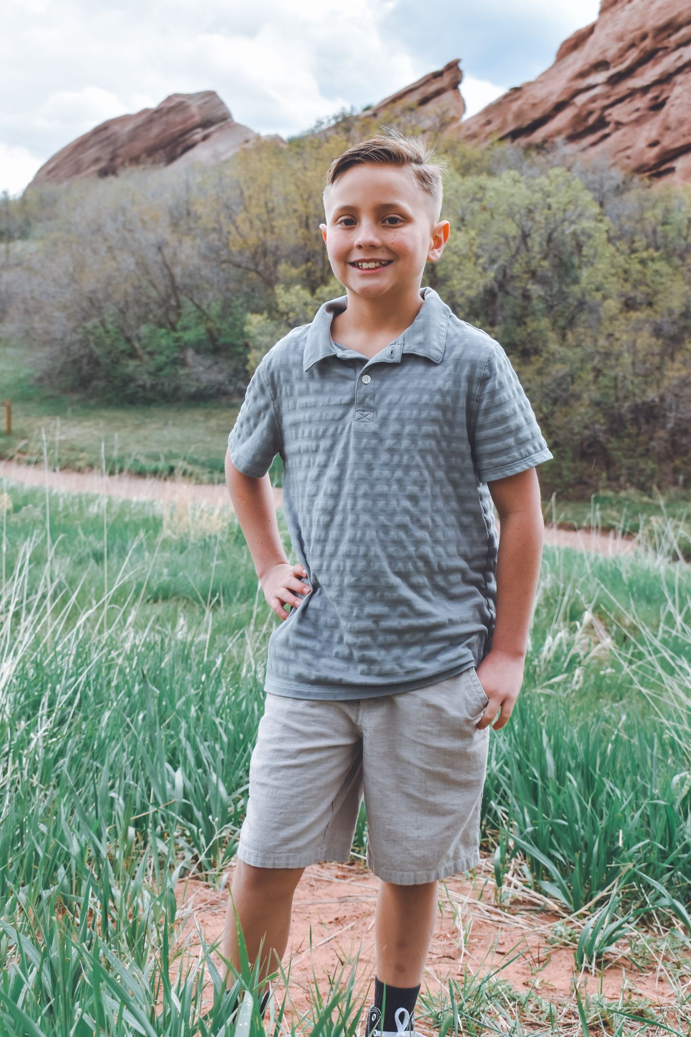 Smiling boy in gray polo shirt and khaki shorts standing in front of red rock formations outdoors. Family Photographer in Pflugerville, Tx