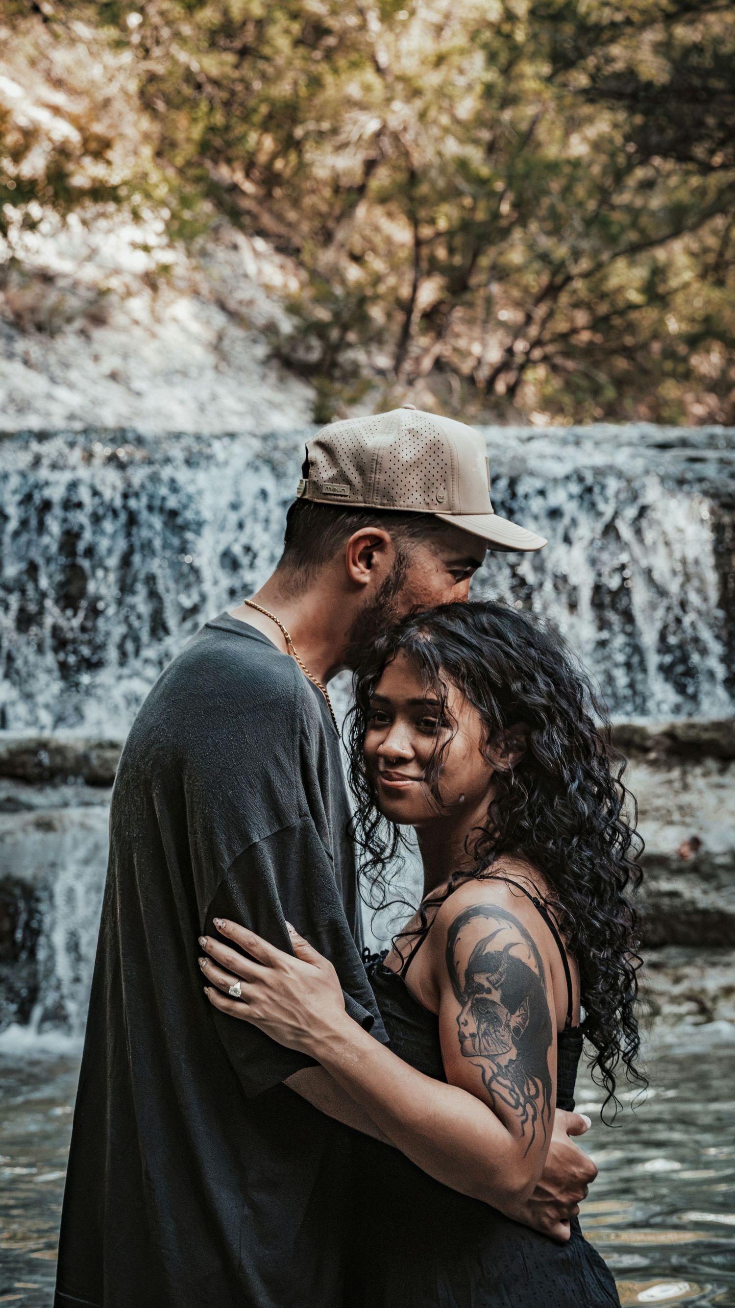 Tattooed couple embracing tenderly near a flowing waterfall in a lush outdoor setting for a couples session.