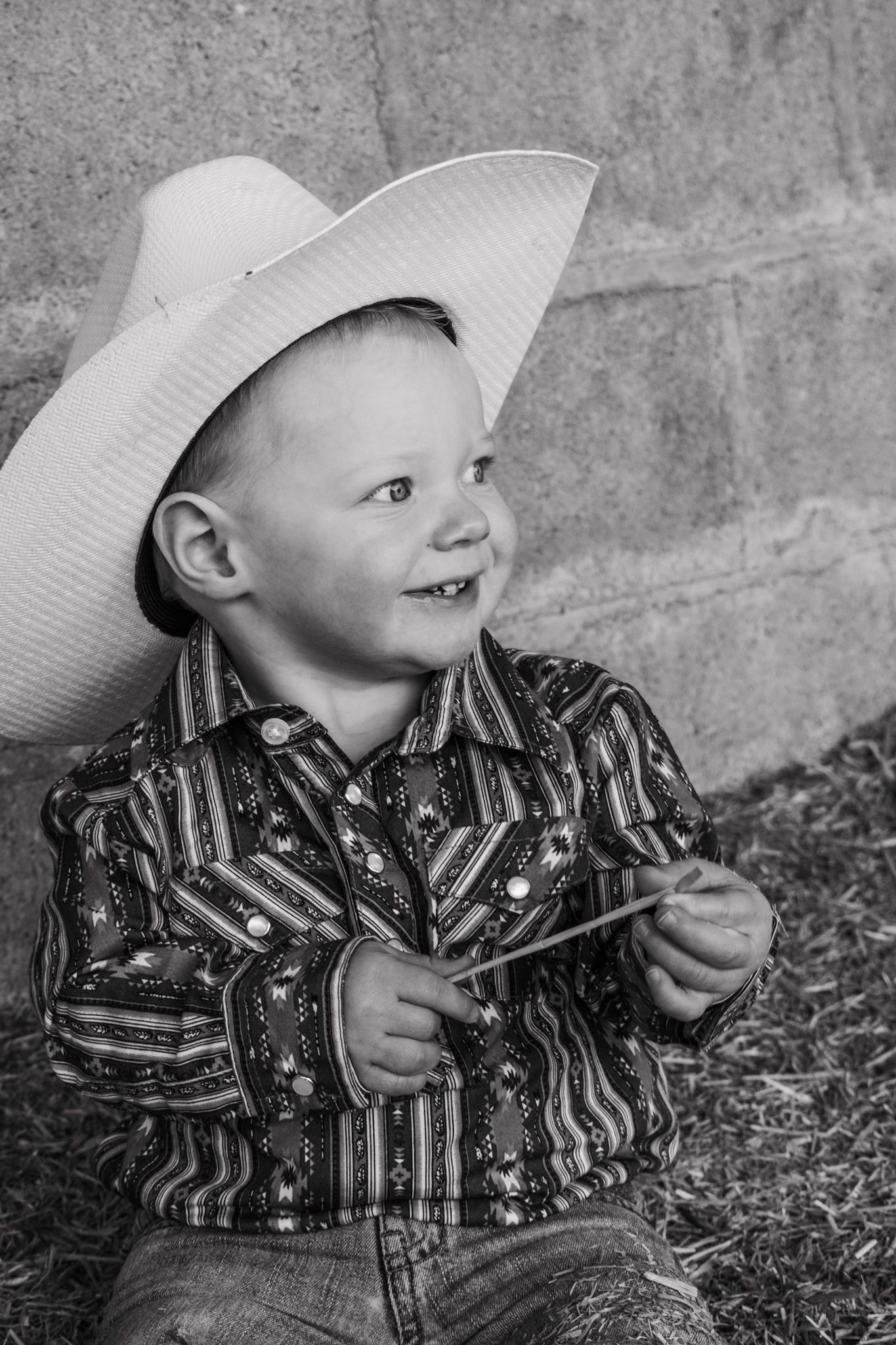 Smiling young boy in cowboy hat and western shirt sitting in hay in black and white photo. Family Photographer in Pflugerville, Tx