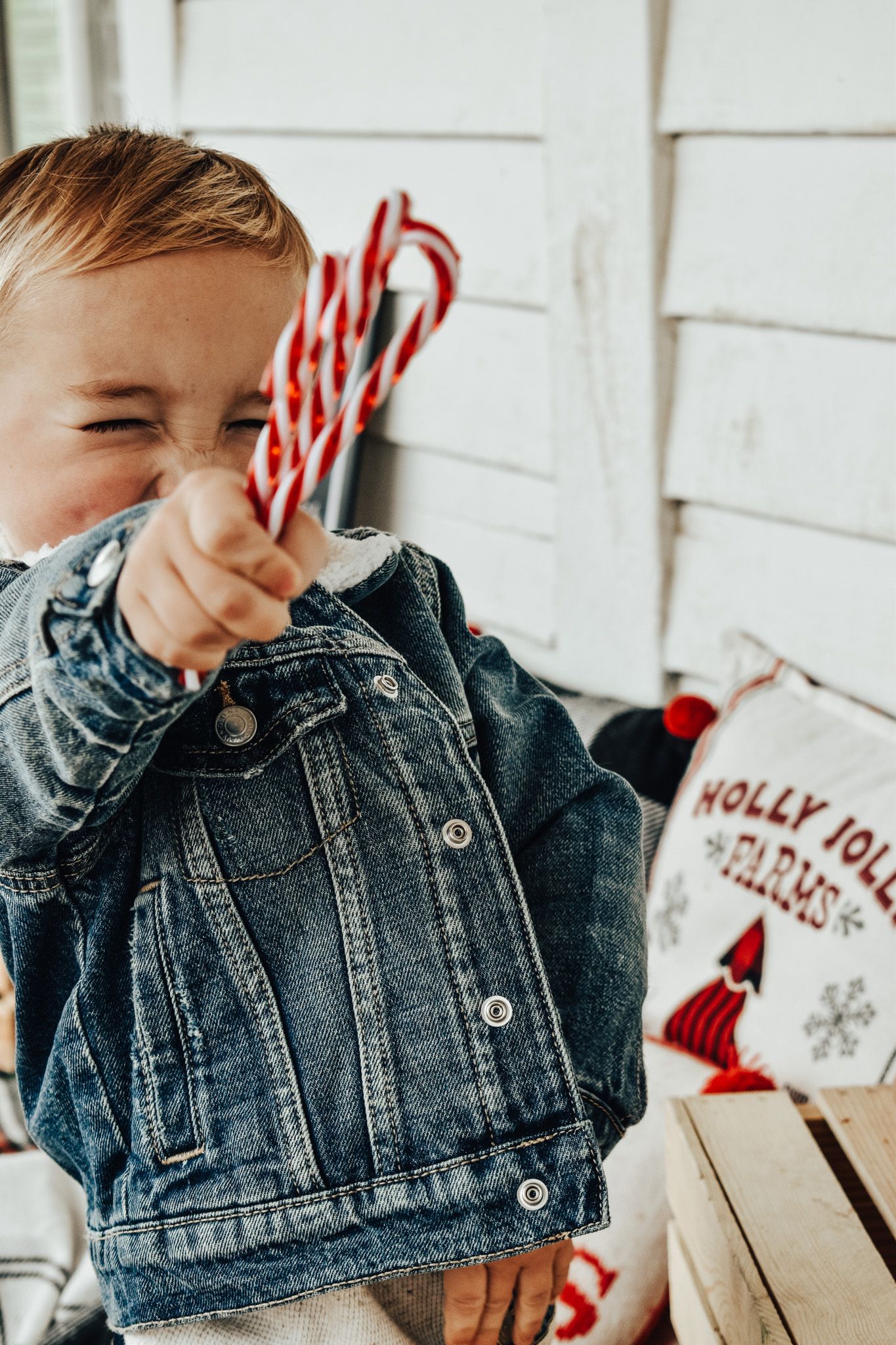 Cheerful toddler in denim jacket holding candy canes on a festive Christmas porch setting.