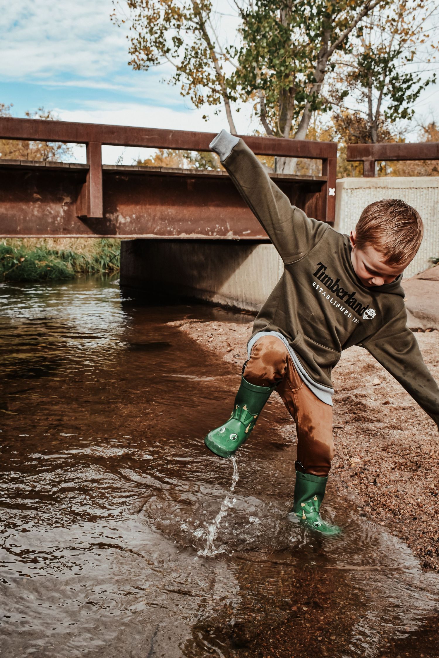 Excited young boy in green rain boots splashing water in a creek near a wooden bridge in autumn.