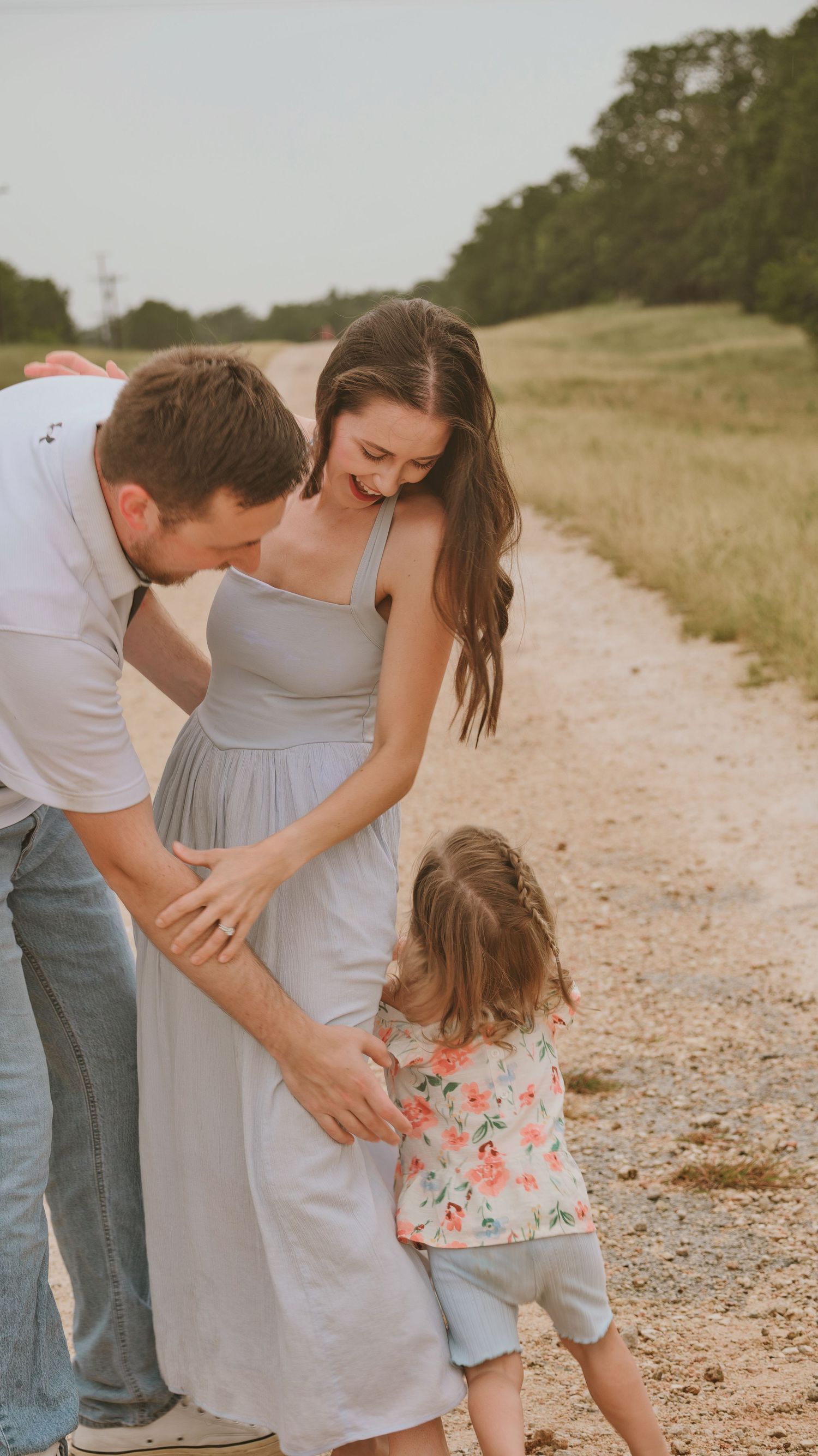 Parents smiling down at their young daughter on a gravel path during a golden hour photo session.