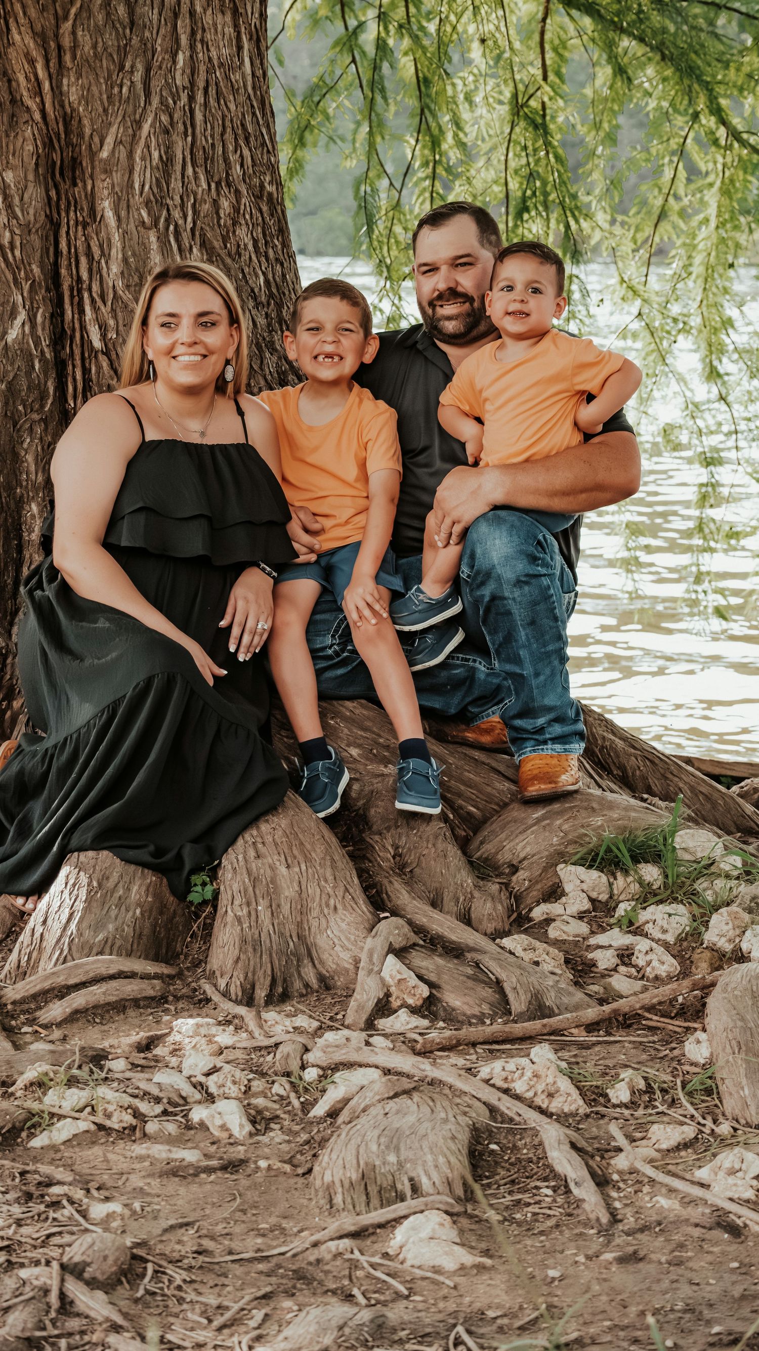 Happy family of four sits on tree roots by a lake, boys wearing matching orange shirts.