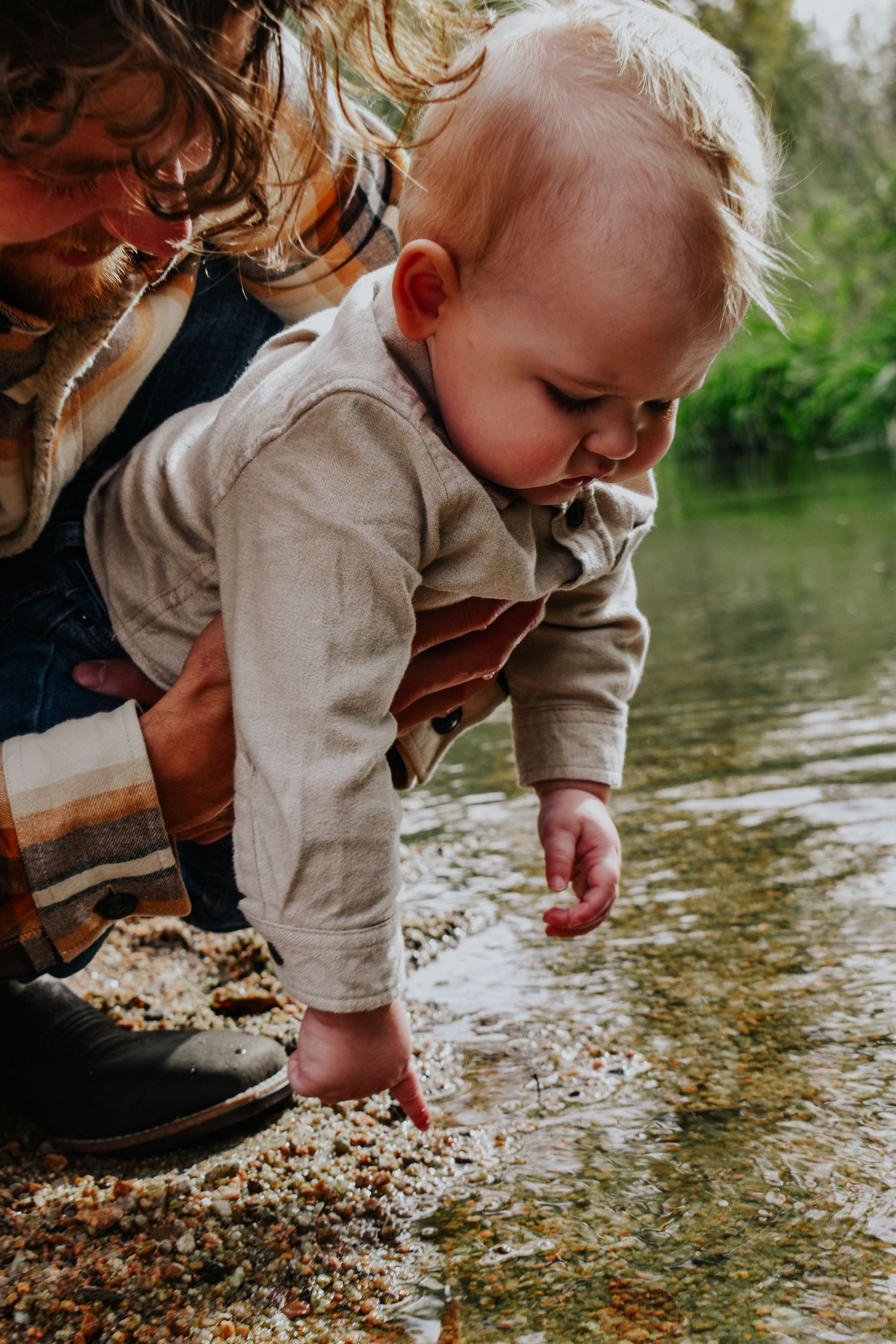 Father supporting baby as curious blonde toddler reaches down to touch shallow river water.