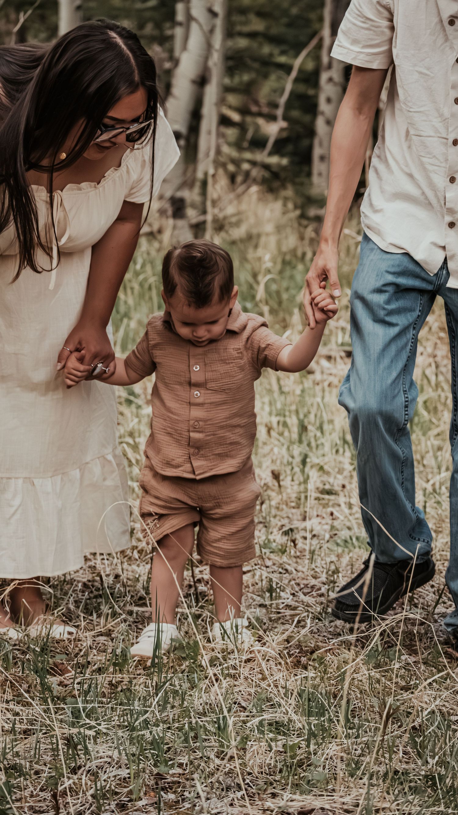 Parents helping toddler boy take first steps through a grassy forest clearing holding both hands.