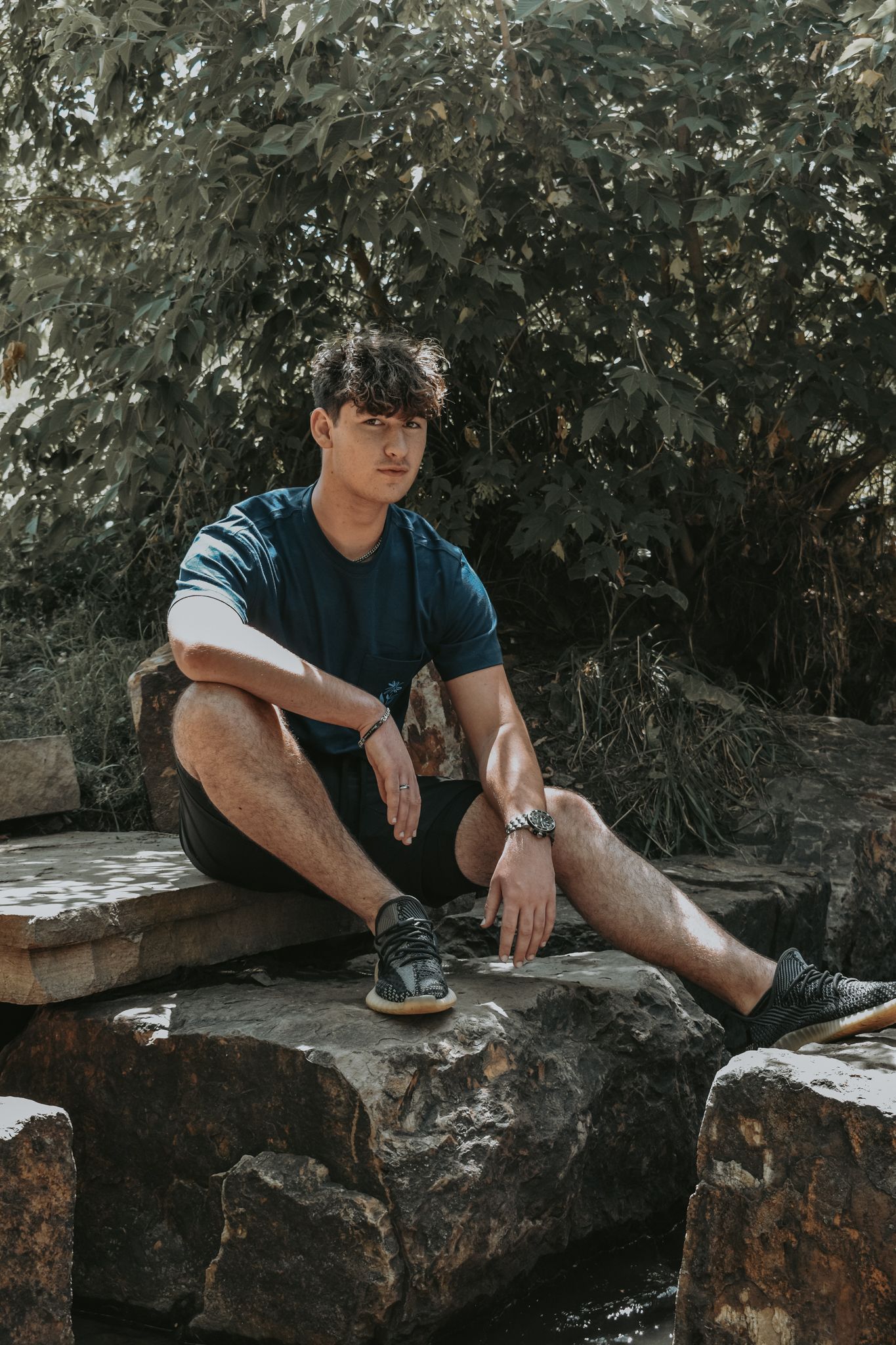 Young man in blue t-shirt sitting on stone steps near a rocky stream with green trees in background for senior photos.