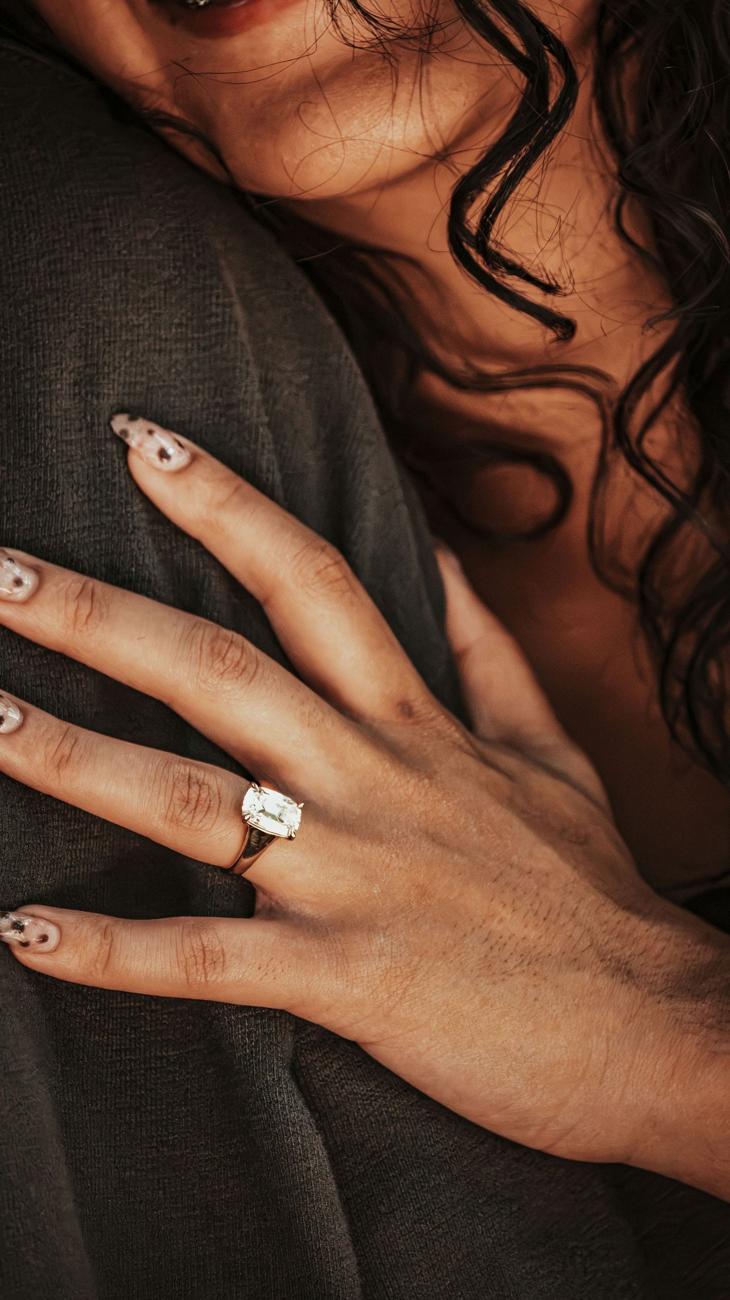 Close-up of womans hand with large emerald-cut engagement ring resting on mans gray shirt sleeve.
