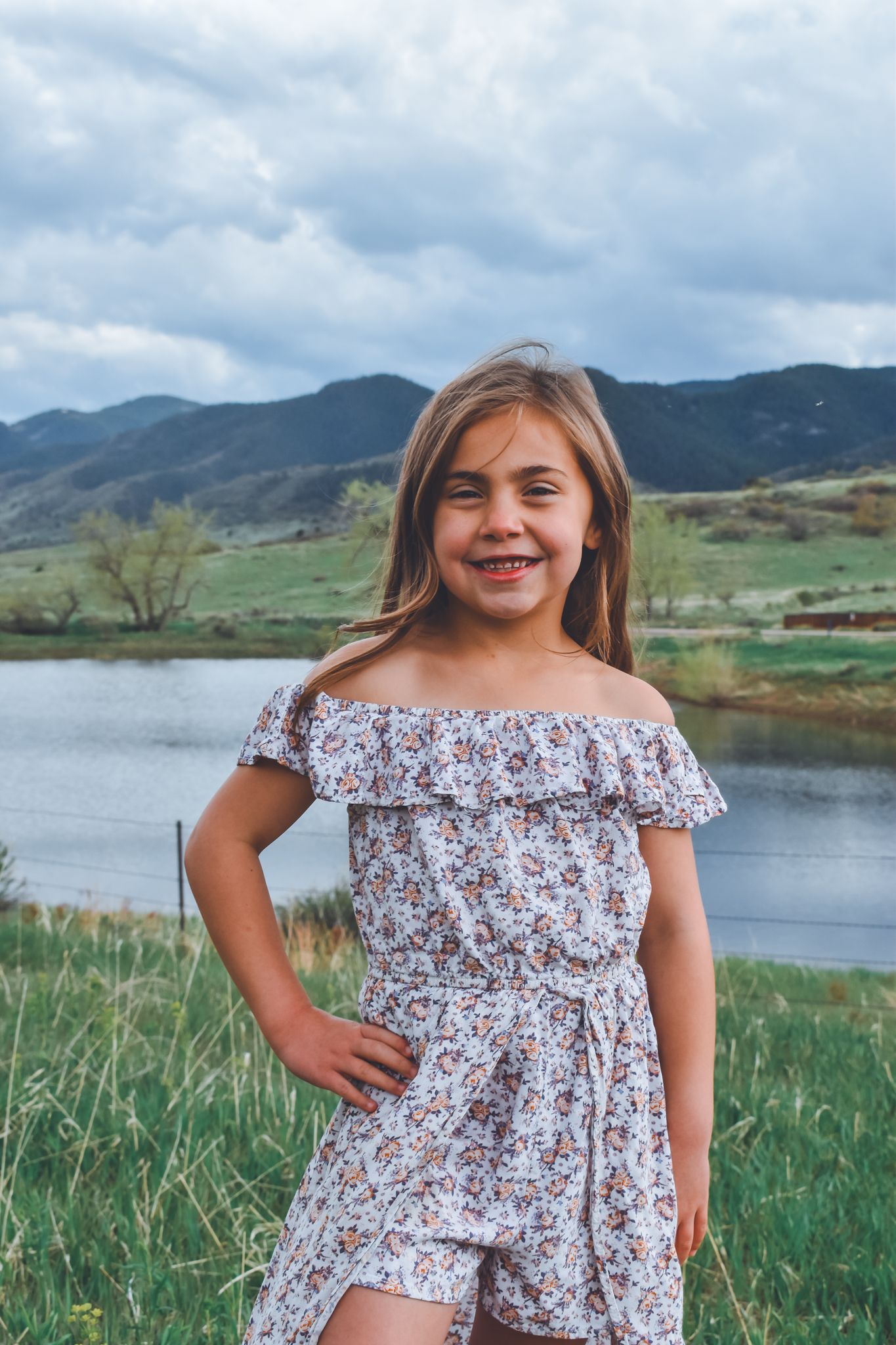 Young girl in floral off-shoulder romper posing near a lake with mountain views in background. Family Photographer in Pflugerville, Tx