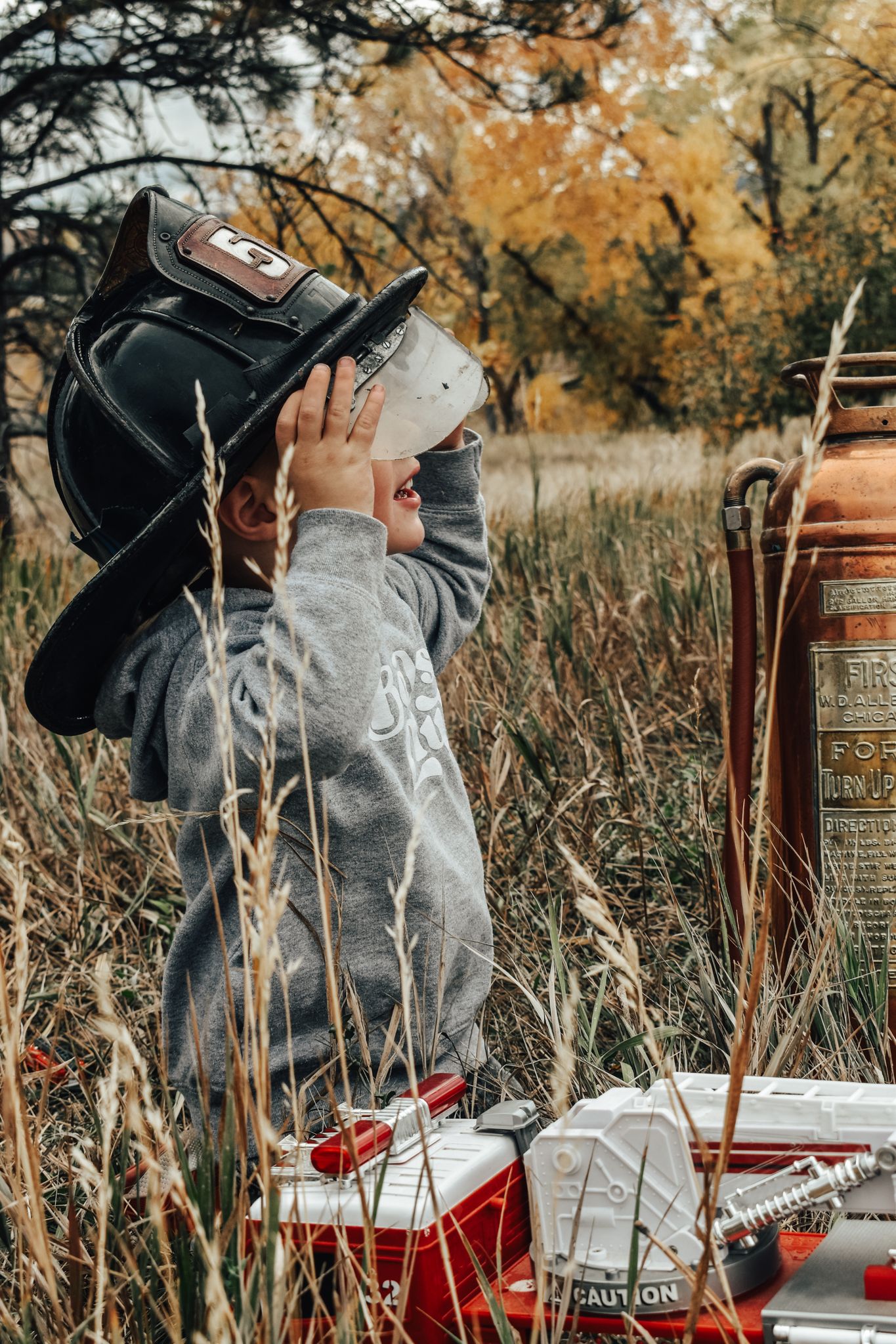 Young child wearing firefighter helmet playing with toy fire truck in tall autumn grass. Family Photographer in Pflugerville, Tx
