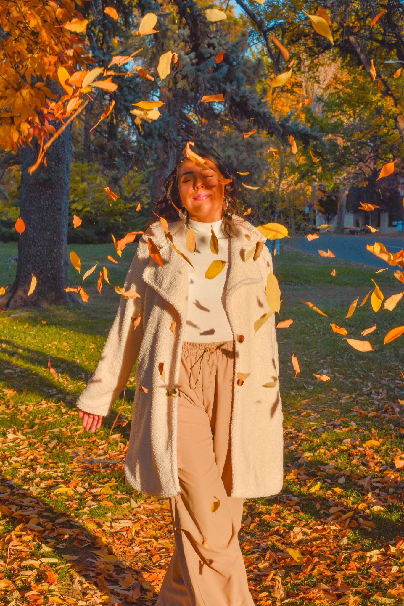 Woman in cream coat smiling as golden autumn leaves fall around her in sunny park for senior photos.