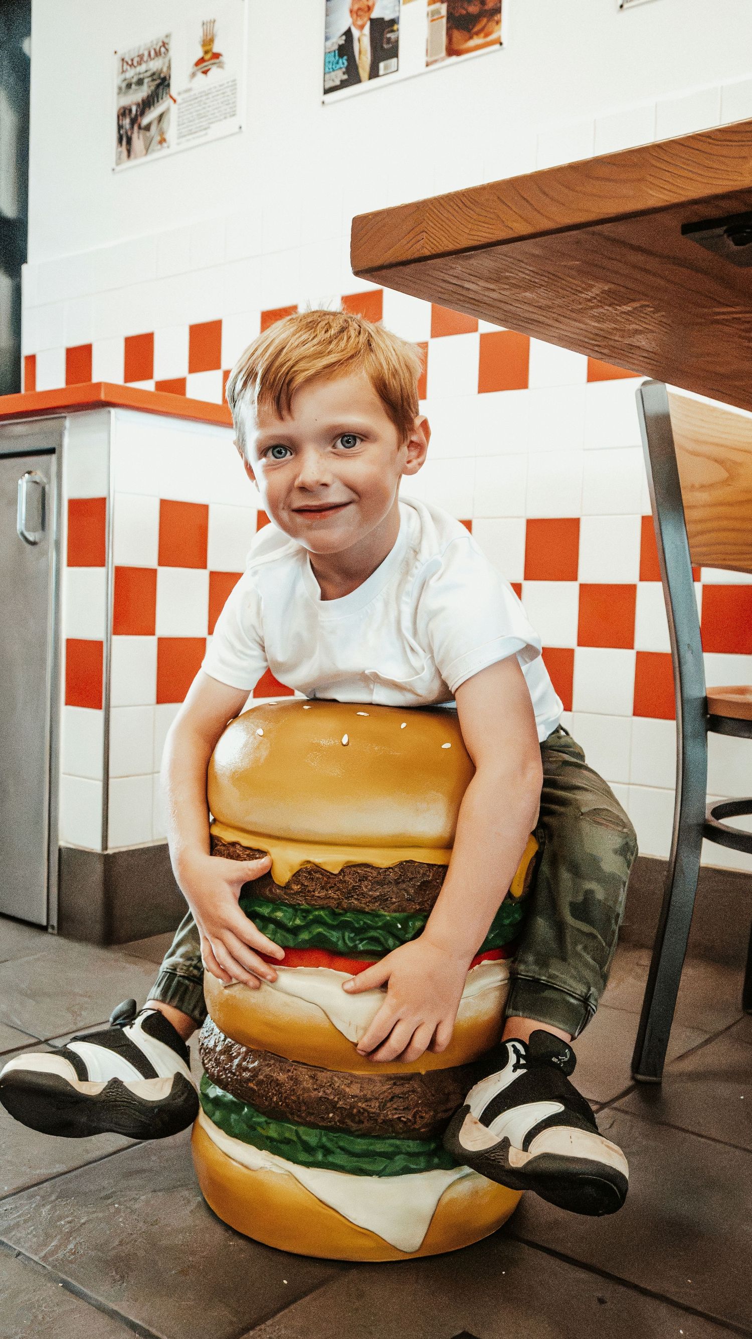 Happy boy hugging a large burger statue inside Five Guys restaurant with checkered walls.