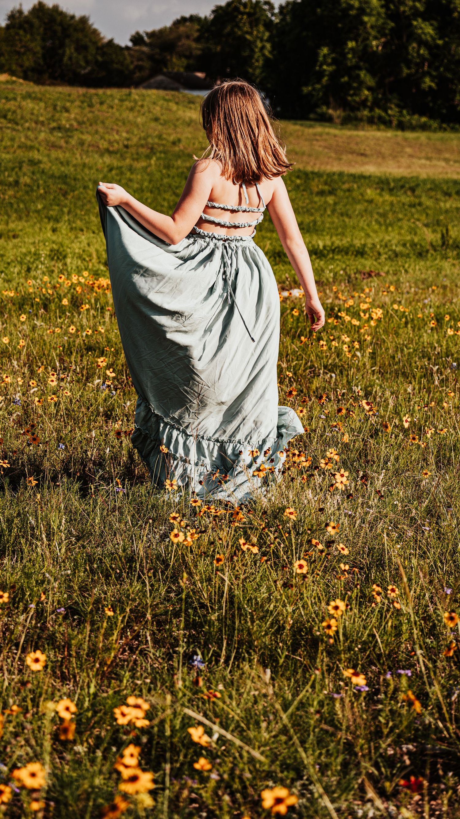 Young girl in sage maxi dress walking through sunny yellow wildflower field rear view.