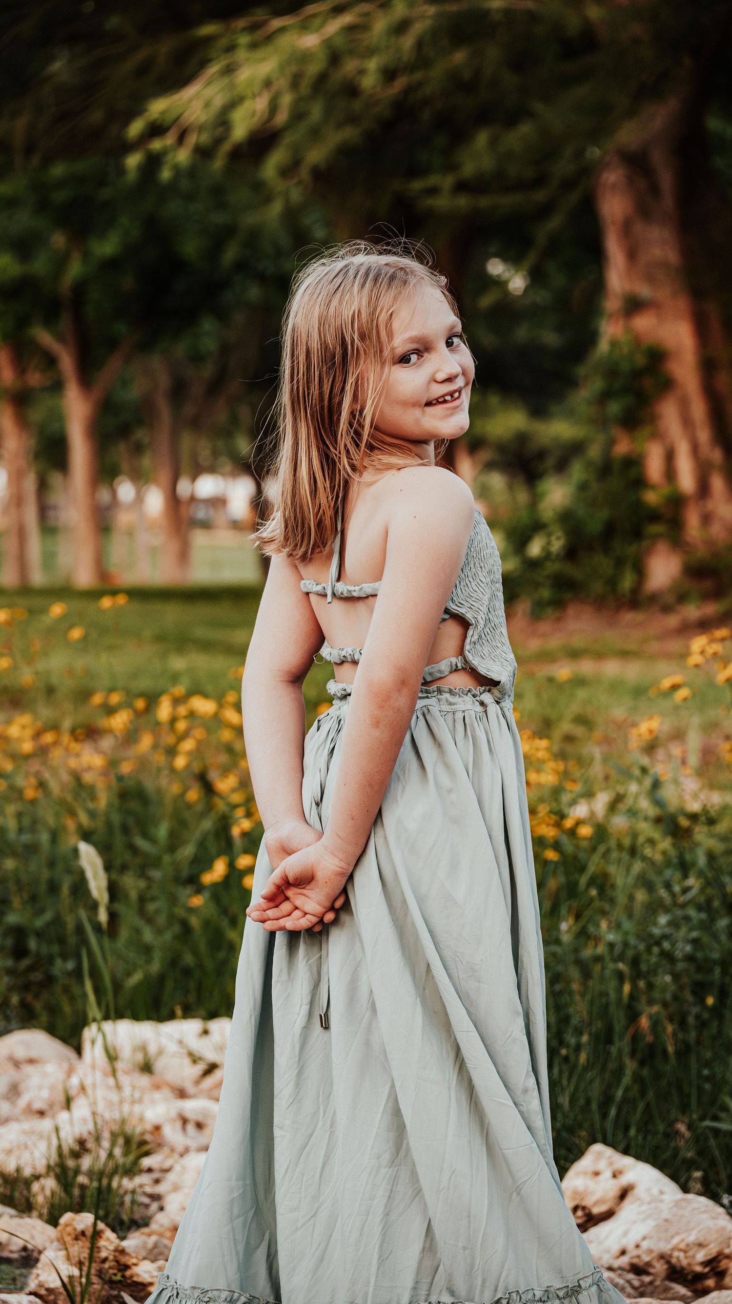 A cheerful young girl in a sage green maxi dress smiles over her shoulder in a sunny garden setting.