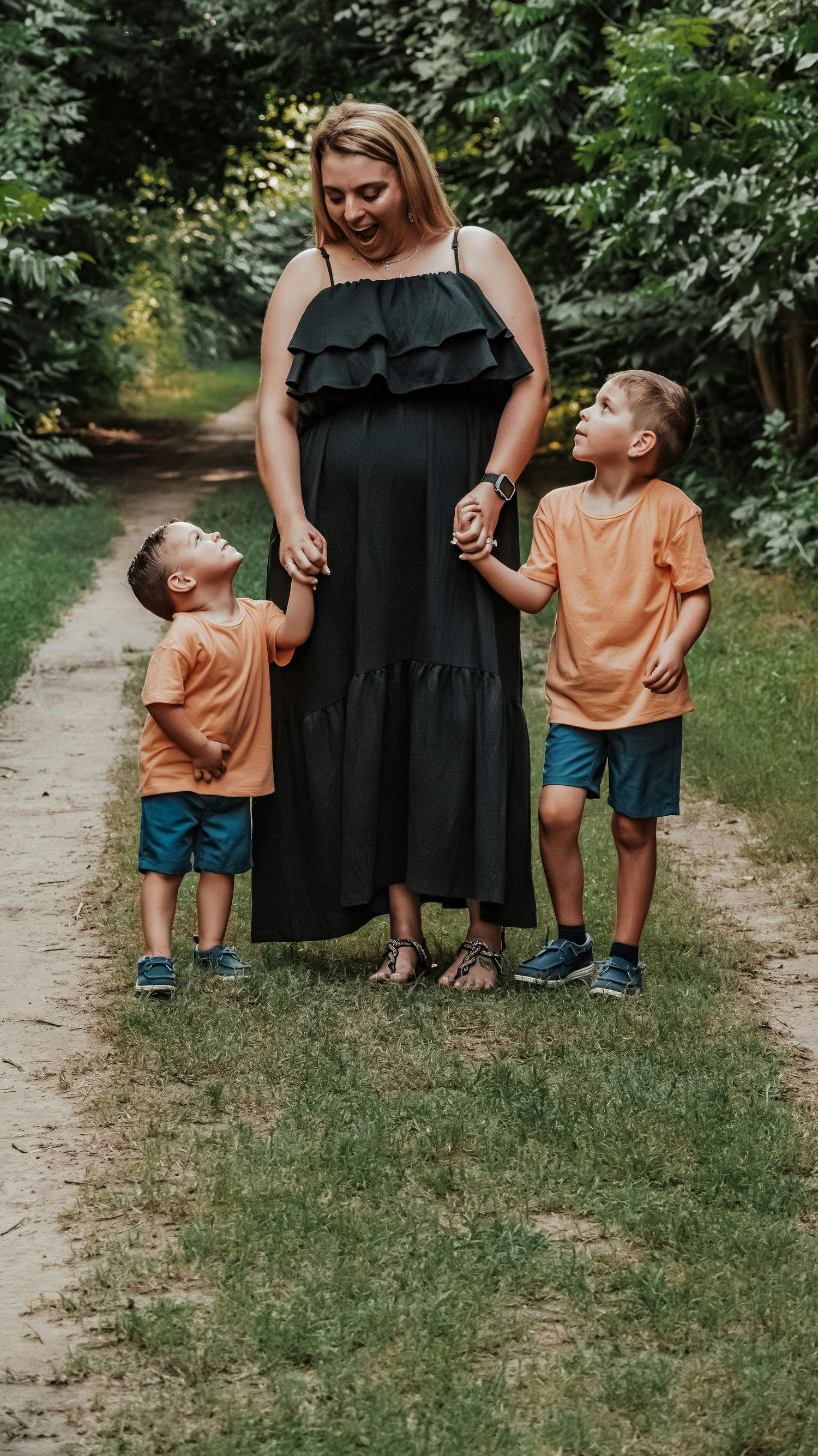 Smiling pregnant mom in black dress walking with two boys in orange shirts on a lush green nature trail.