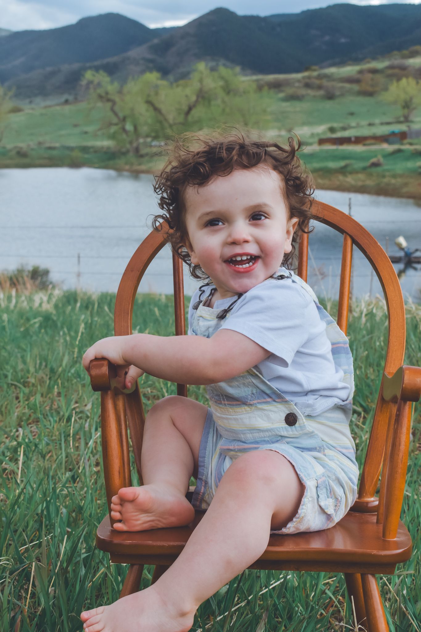 Laughing toddler with curly hair sitting in wooden chair outdoors with mountain lake backdrop.