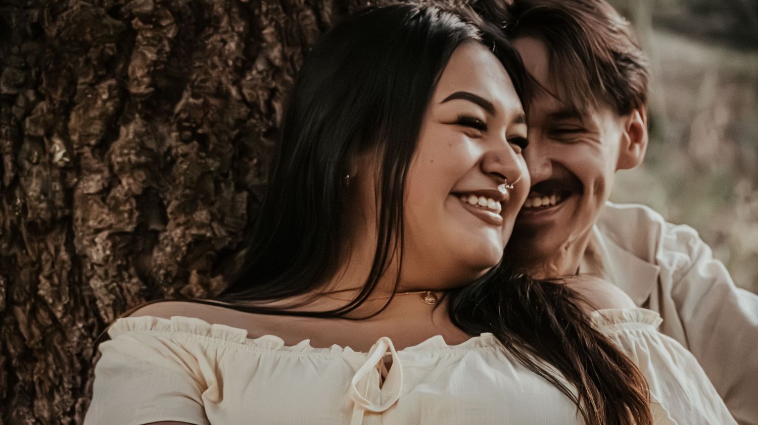 A smiling couple shares a tender moment together, leaning close while seated against a large tree.
