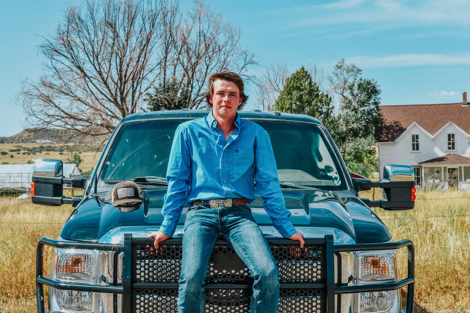 Young man in blue western shirt sitting on hood of green pickup truck in rural farm setting for senior photos.