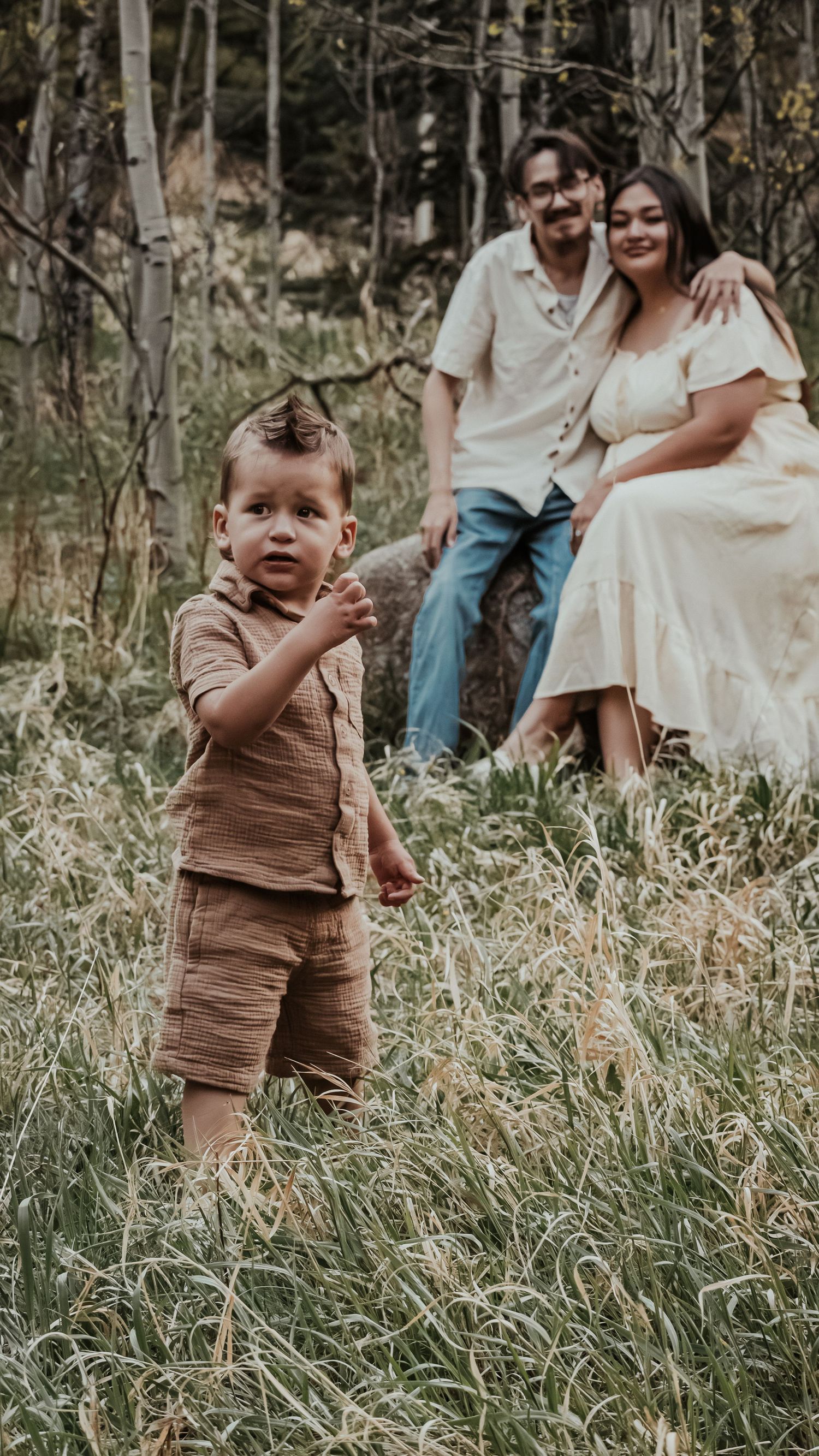 A curious toddler boy in brown shorts explores a grassy meadow while his parents relax behind him.