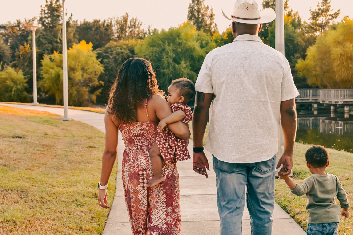 A Black family walks along a park path at sunset, mother carrying baby girl, father holding young son's hand.