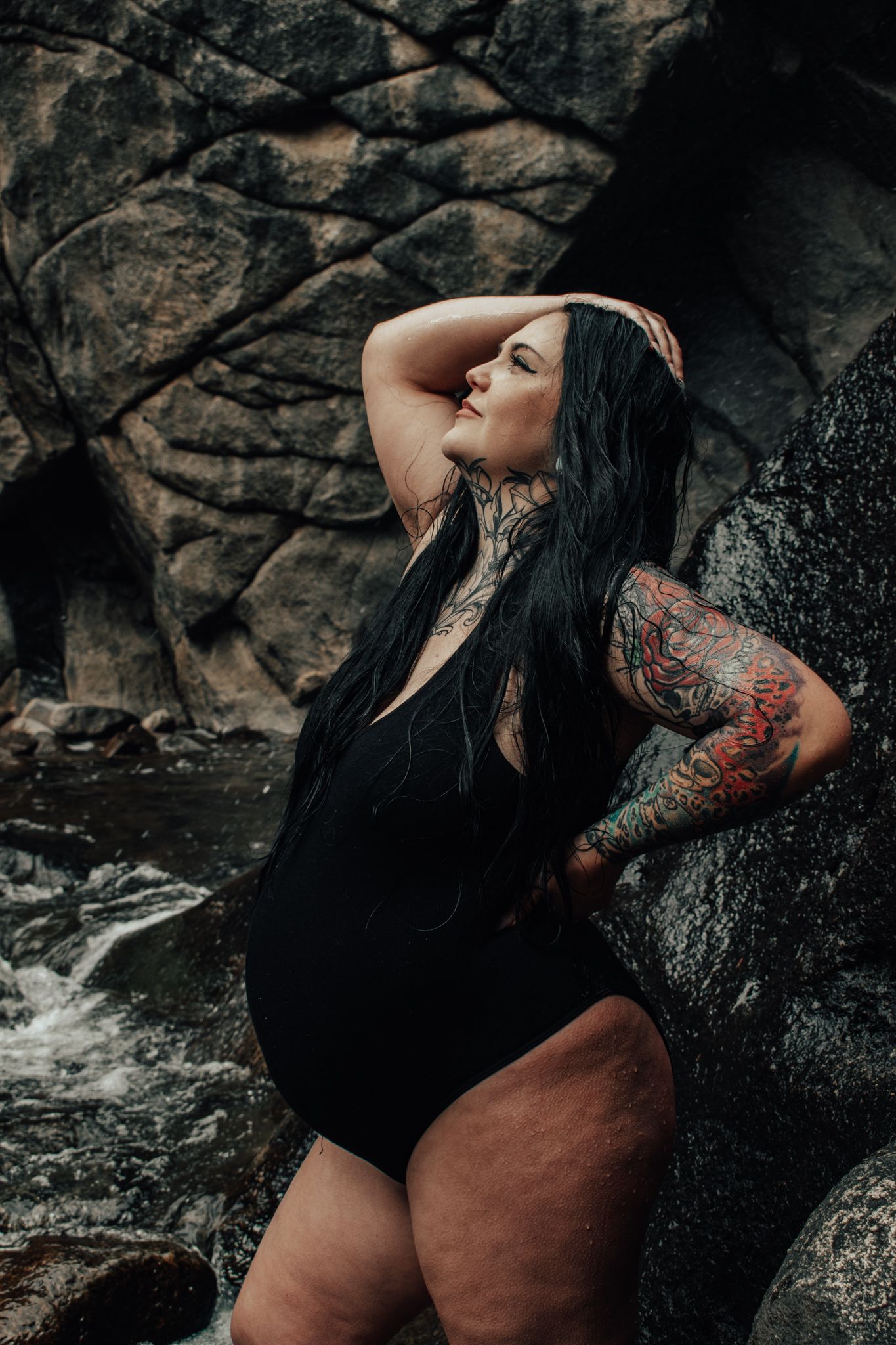 Tattooed woman in black swimsuit posing confidently against rocky waterfall backdrop.