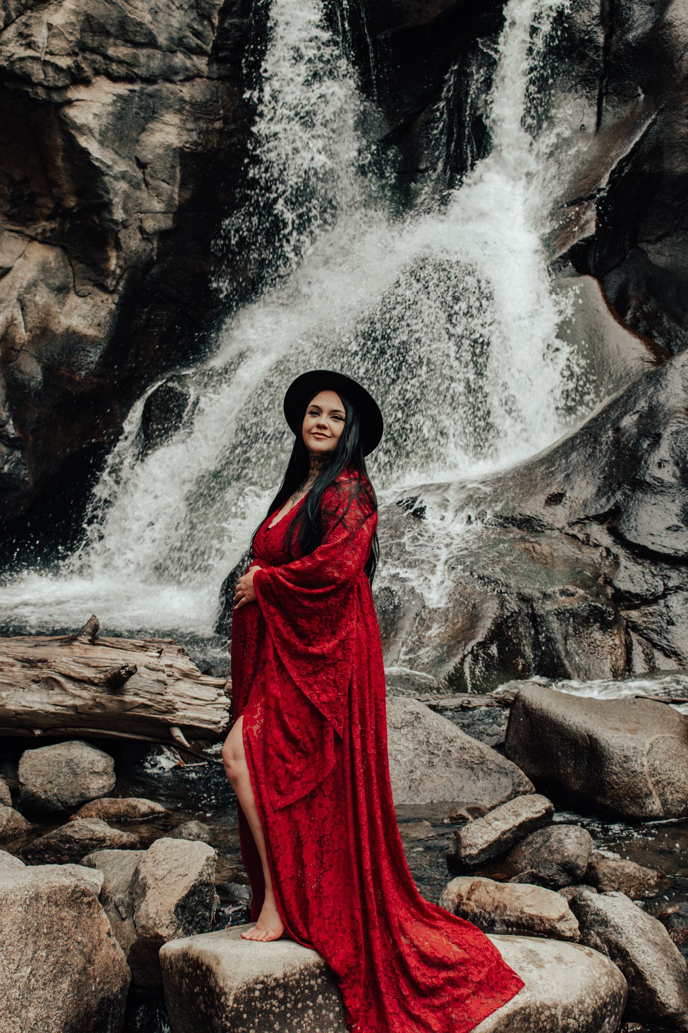 Pregnant woman in flowing red lace gown and black hat stands on rocks before waterfall.