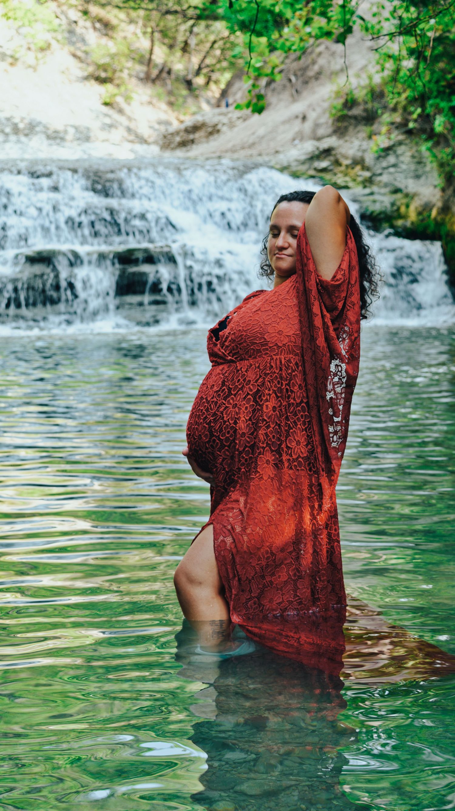Pregnant woman in red lace dress standing in turquoise water before a cascading waterfall.