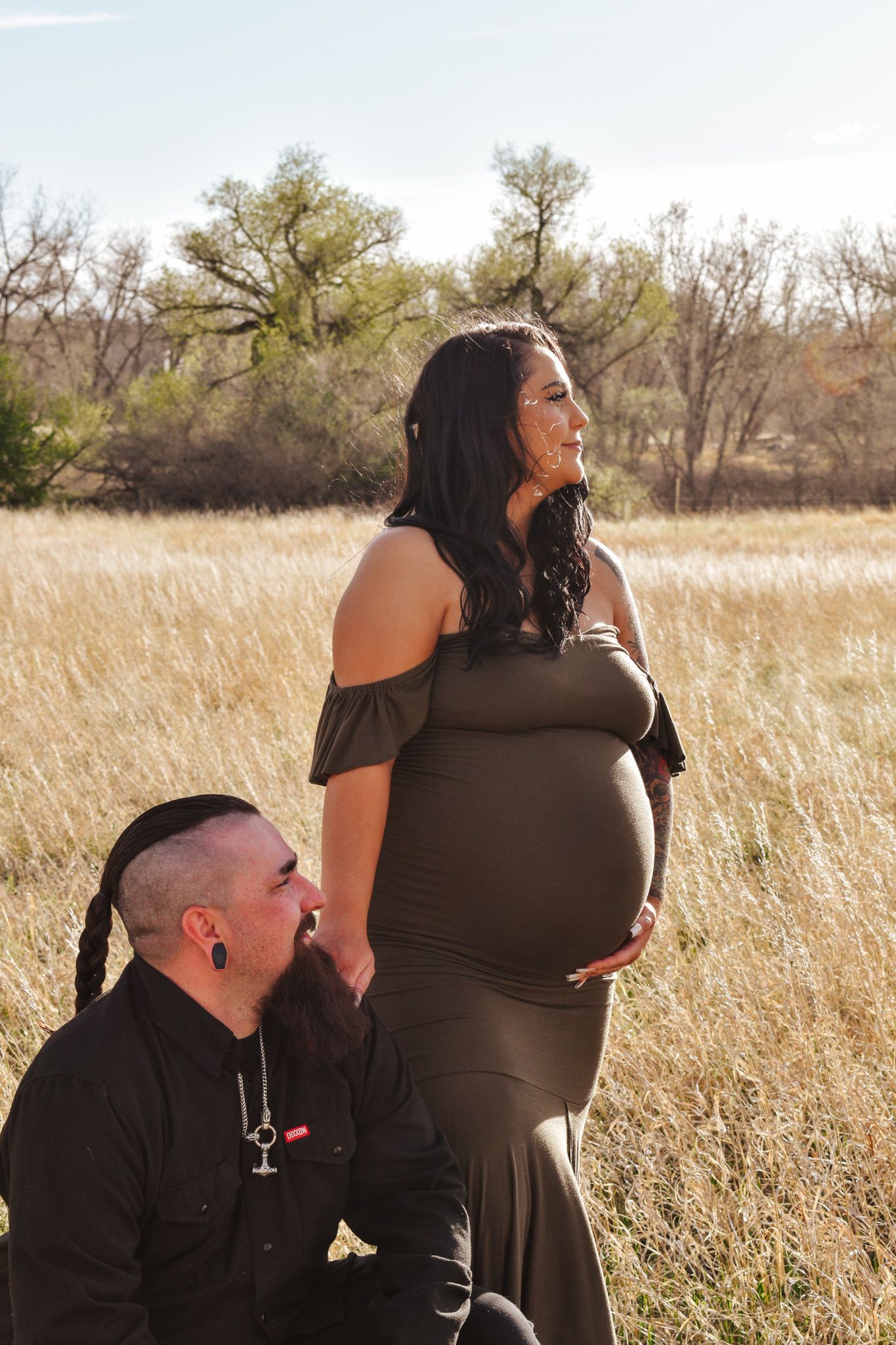 Pregnant woman in olive mermaid dress cradles baby bump while partner kneels beside her in sun-drenched autumn field.