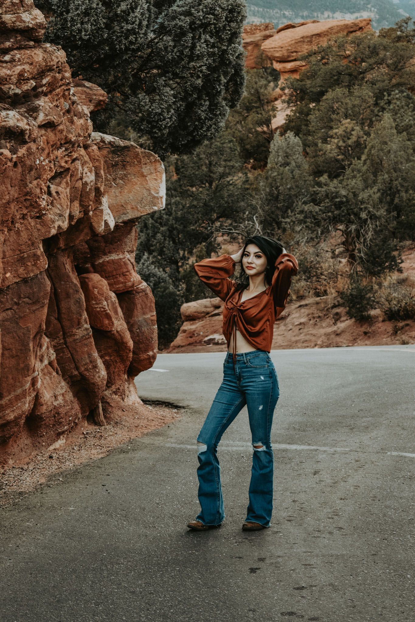 Stylish young woman in rust top and flare jeans posing by red rock formations.