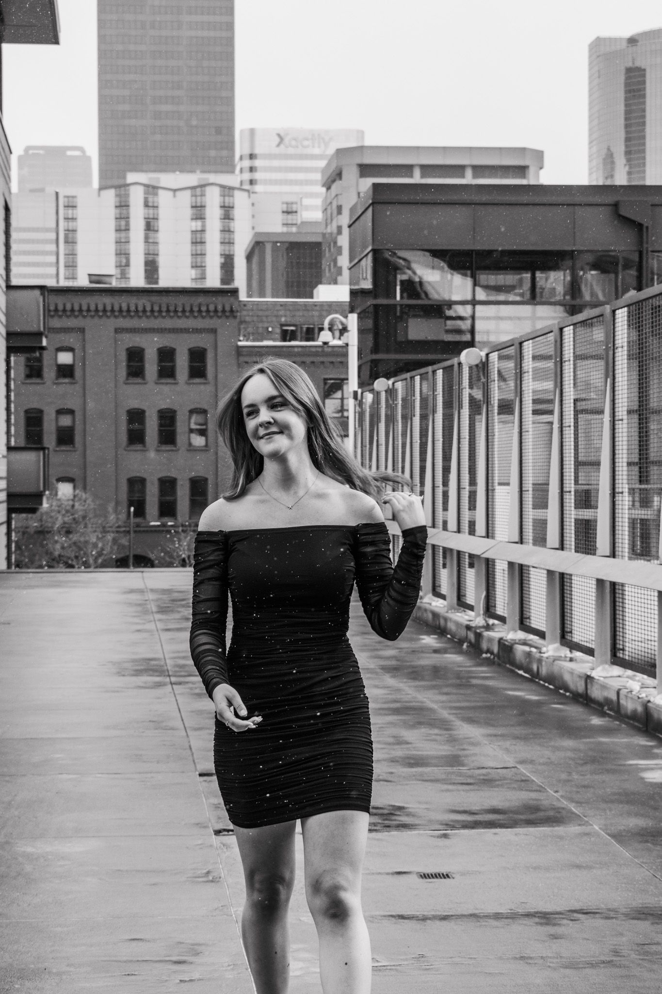 Young woman in black off-shoulder mini dress walking on urban rooftop with city skyline, black and white photo for senior photos.