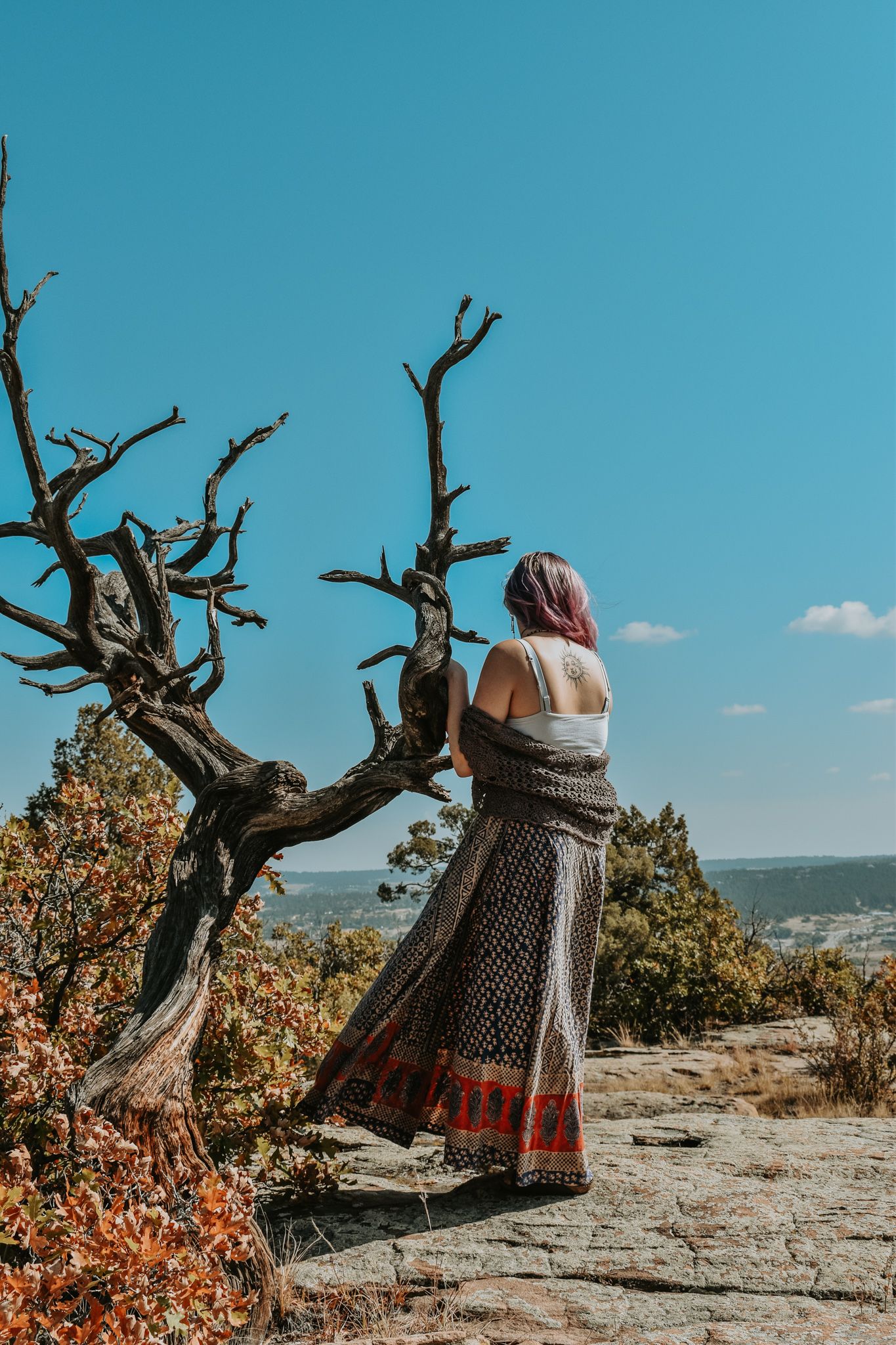 Woman with pink hair in boho skirt stands beside gnarled dead tree on rocky overlook for senior photos.