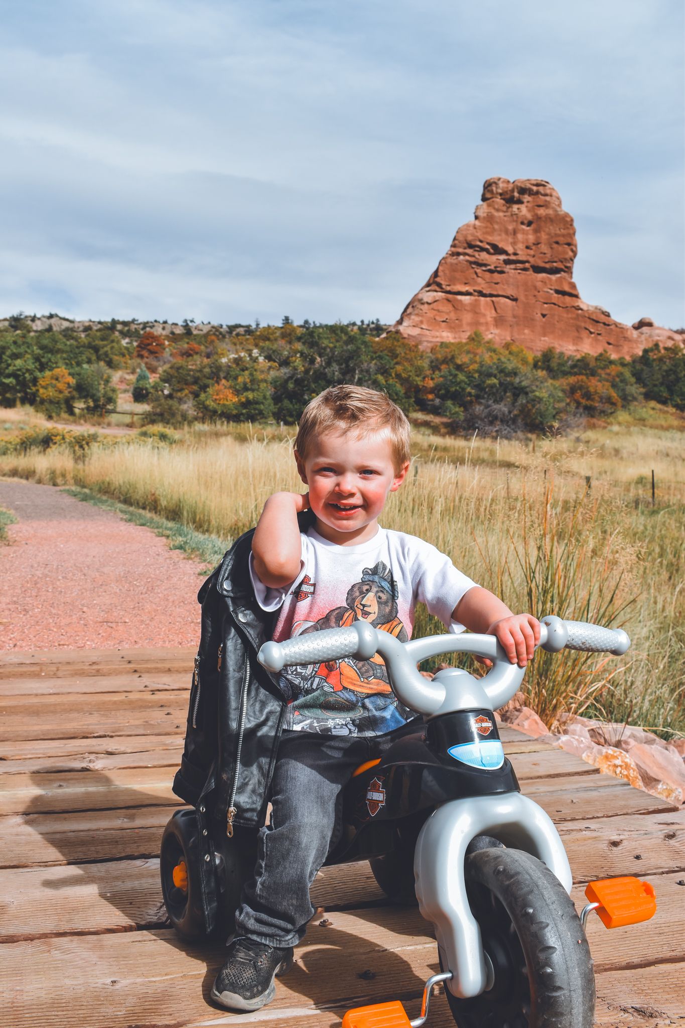 Smiling toddler on toy Harley Davidson tricycle with red rock butte and golden fields in background. Family Photographer in Pflugerville, Tx