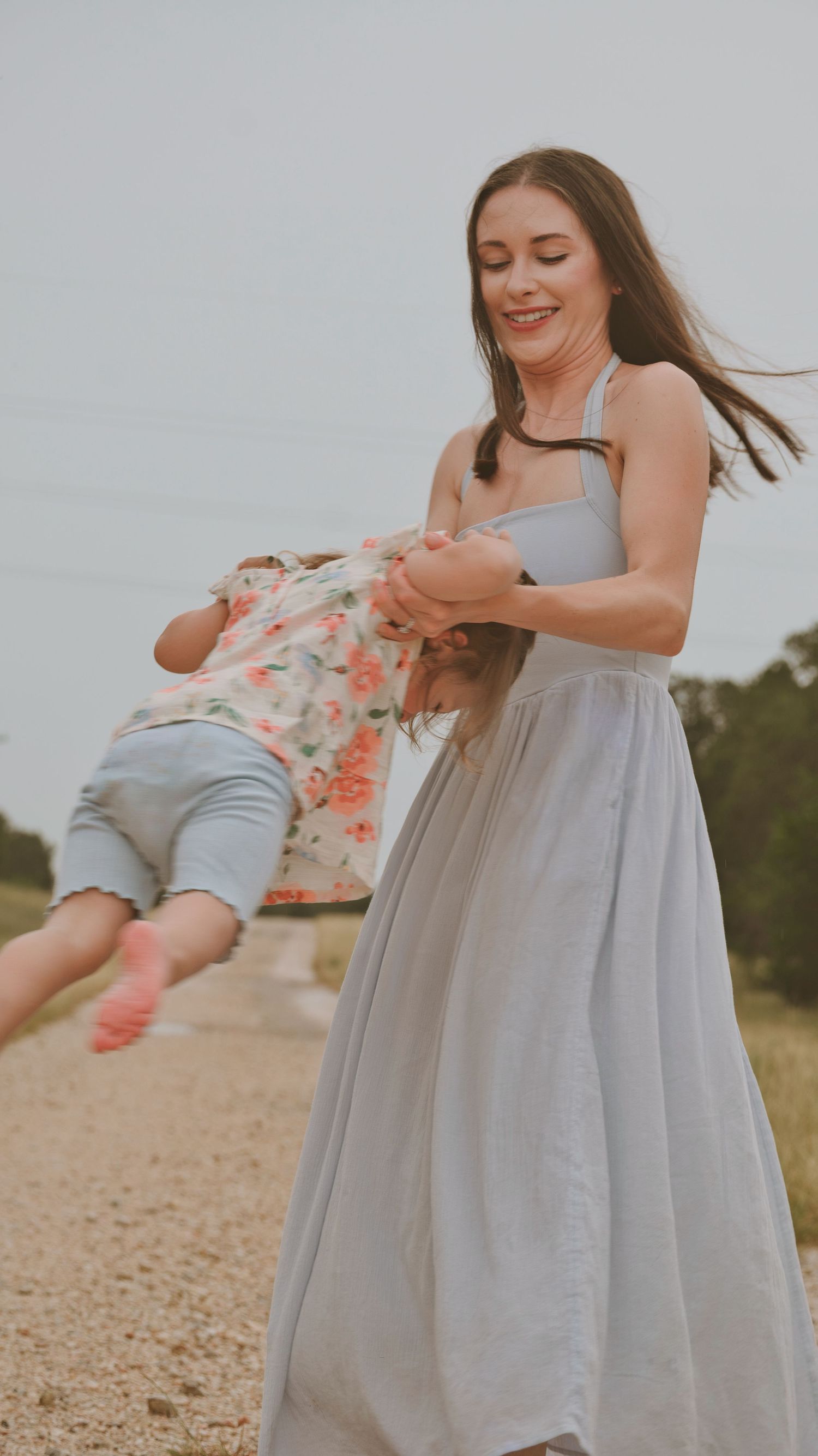 Smiling mother in gray maxi dress spinning her young child on a dirt road outdoors for a family session.