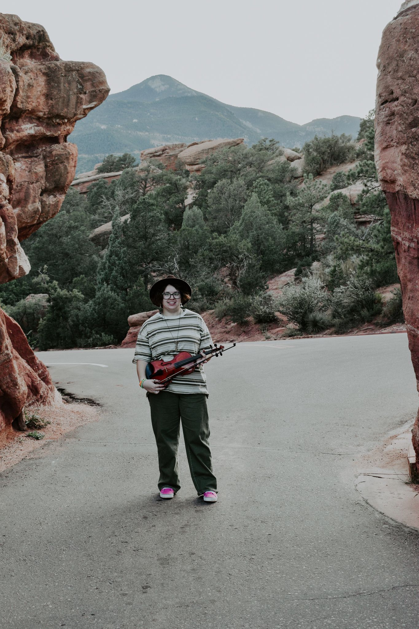 Person standing on a road between red rock formations holding a violin, wearing a striped shirt and hat for senior photos.