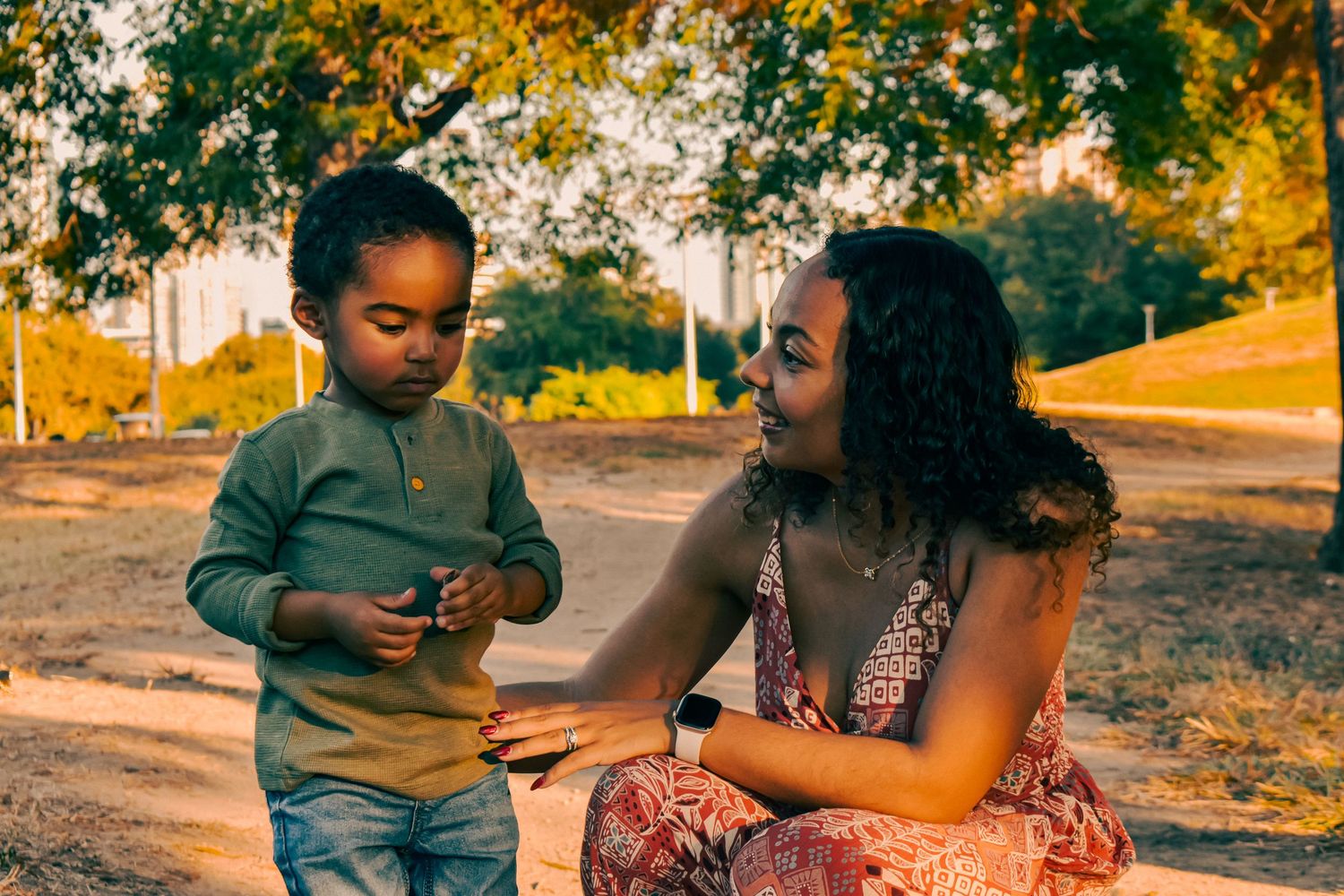 Mother in floral dress crouching to interact with young toddler son in warm golden outdoor park.
