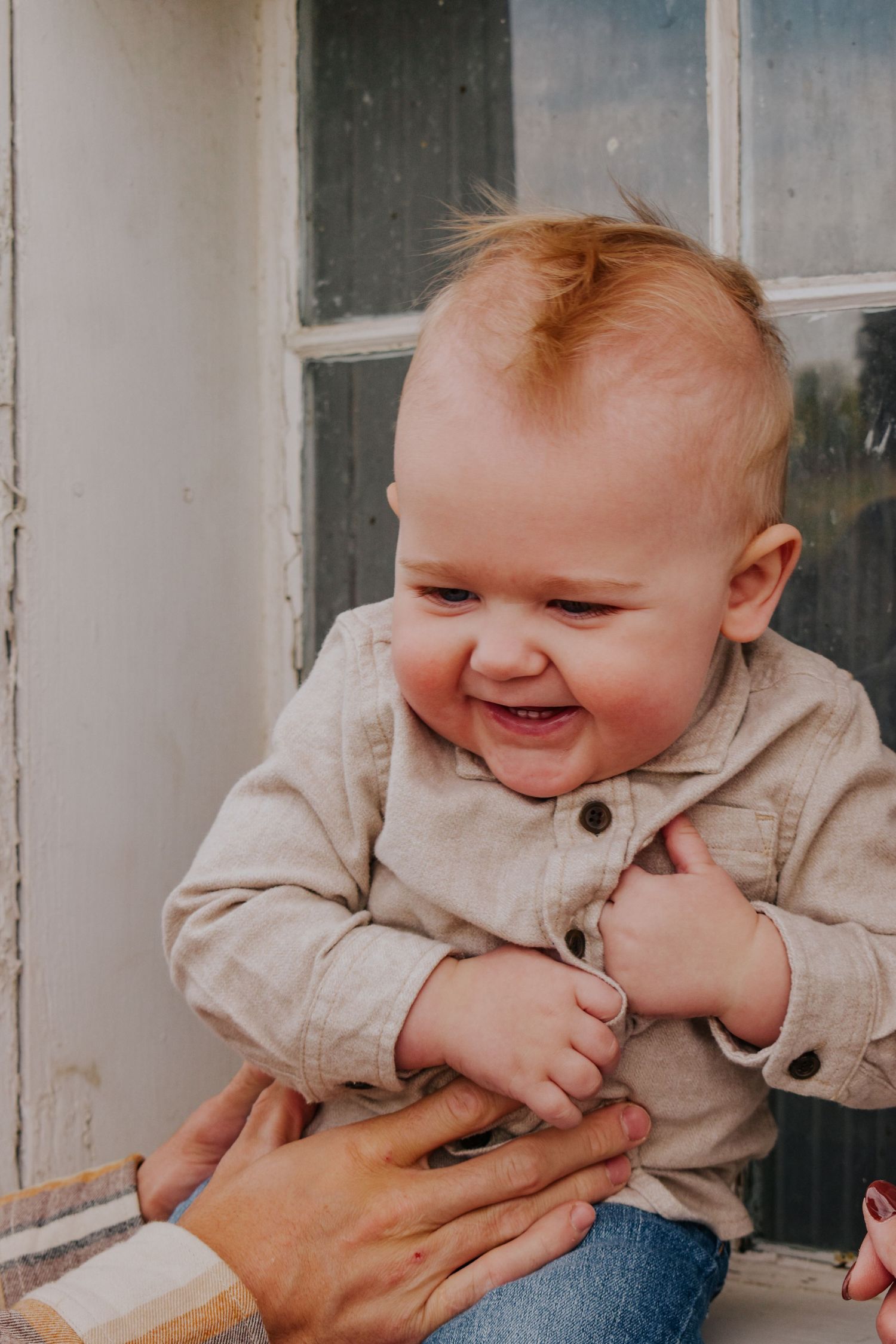 Laughing baby in beige button shirt being held near a vintage window.