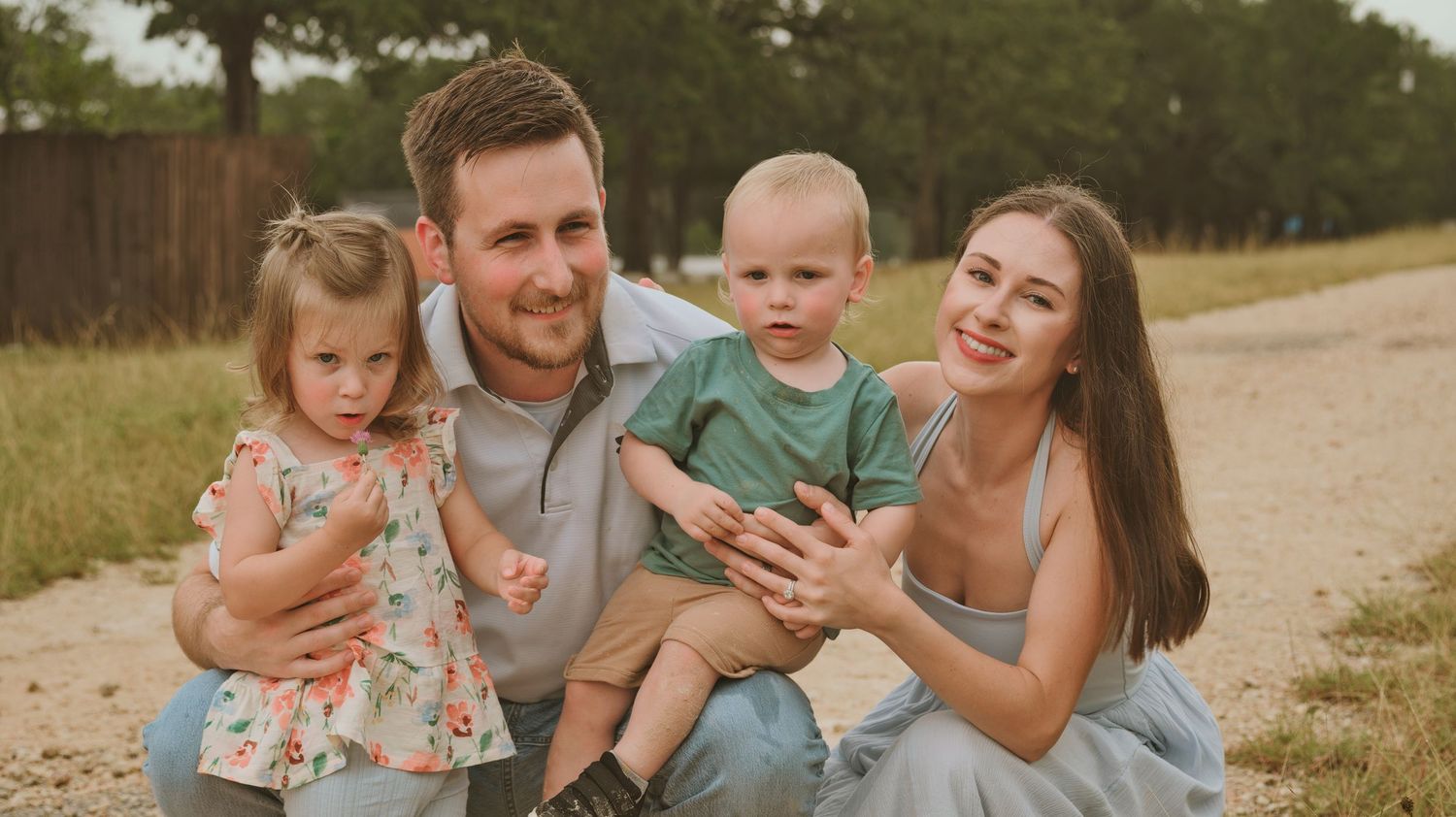 Smiling family of four poses together on a sunny gravel path with trees behind them.