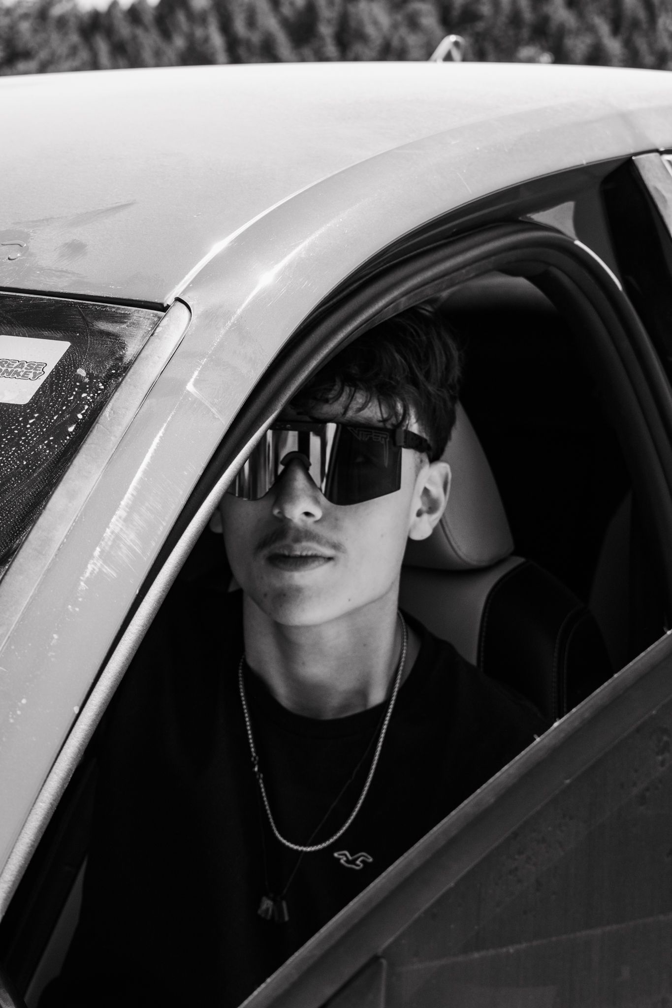 Black and white portrait of a young man wearing sunglasses sitting inside a white car for senior photos.