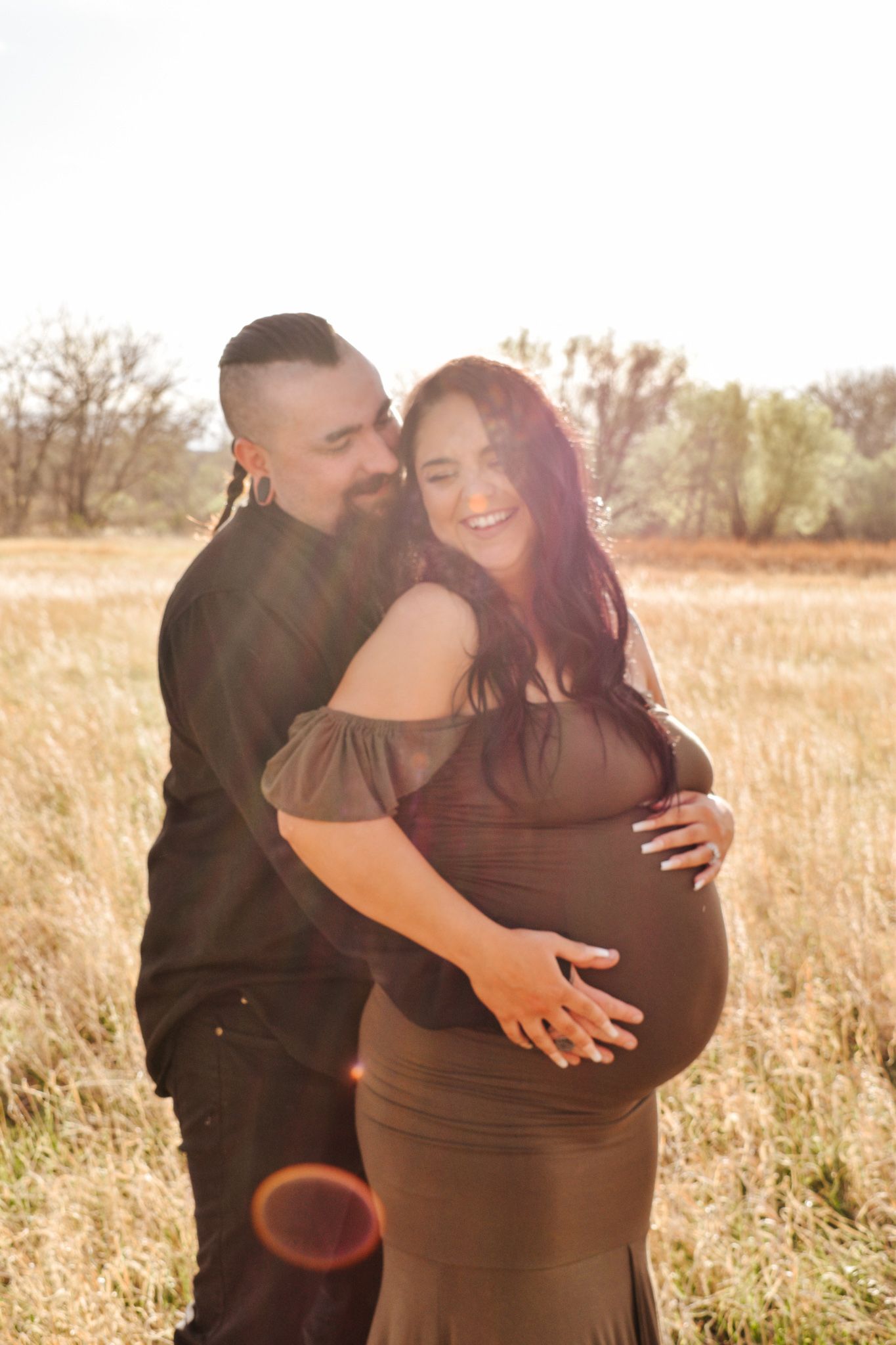 Smiling pregnant couple in sunlit field, man embracing woman in olive maternity gown.